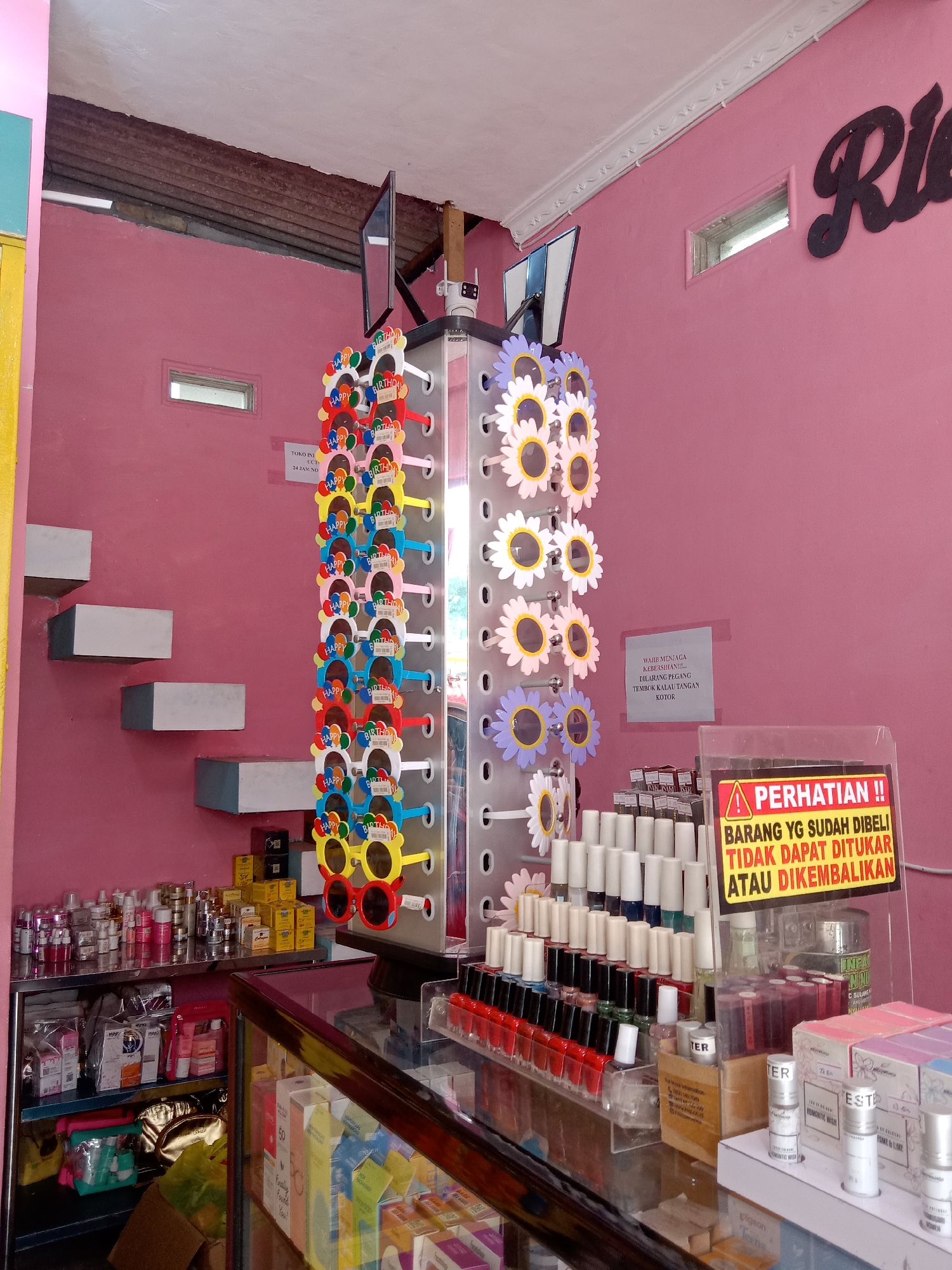 The image depicts the interior of a small retail shop, likely a cosmetic store, located in Sugihwaras, Indonesia. The walls are painted a bright pink, and the ceiling appears to be made of corrugated metal. A black cursive sign with the word "Ria" is affixed to the right wall.
In the foreground, a glass display counter showcases rows of nail polish bottles in various colors. There are also several smaller cylindrical bottles and boxes of cosmetic products. To the left of the counter, shelves are stocked with more beauty products, including lotions and perfumes. In the middle of the image, a rotating display stand features an assortment of novelty sunglasses shaped like sunflowers and with "Happy Birthday" inscribed on them. The display stand also has a small security camera mounted at the top.
A sign is prominently displayed near the sunglasses, with the text "PERHATIAN!! BARANG YG SUDAH DIBELI TIDAK DAPAT DITUKAR ATAU DIKEMBALIKAN" which translates to "ATTENTION!! GOODS THAT HAVE BEEN BOUGHT CANNOT BE EXCHANGED OR RETURNED". Another smaller sign on the pink wall reads "WAJIB MENJAGA KEBERSIHAN... DILARANG PEGANG TEMBOK KOTOR" meaning "MUST MAINTAIN CLEANLINESS... DO NOT TOUCH THE DIRTY WALL".
To the left of the display stand, a series of grey rectangular shelves are built into the pink wall, resembling steps. The overall impression is that of a brightly colored, compact retail environment selling beauty products and novelty items. There are no people visible in the image.