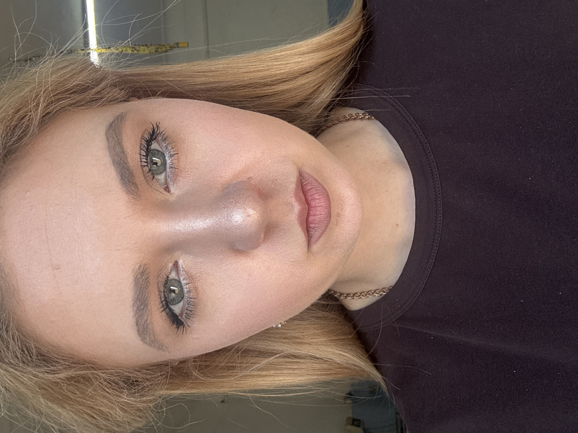 A close-up, head-and-shoulders portrait shows a young woman with blonde hair and green eyes. She is wearing a dark purple shirt and a gold chain necklace. Her makeup includes mascara and lipstick. The background is blurred and appears to be an indoor setting with fluorescent lighting. The image is not detailed enough to provide specific location information.