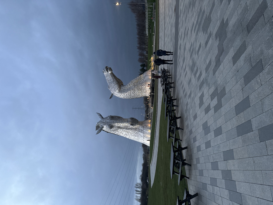 The image displays an outdoor scene at The Kelpies, located in The Helix park, Falkirk, United Kingdom, under an overcast, dark grey sky. Two monumental, metallic horse-head sculptures, illuminated from below, dominate the mid-ground. The sculpture on the left faces forward, while the one on the right has its head raised and turned. In the foreground, a paved area with grey and darker grey rectangular pavers features a linear arrangement of at least seven dark-colored wooden picnic tables with attached benches. Four individuals are walking away from the viewer towards the sculptures in the mid-ground; two in dark jackets are in the foreground of this group, with two others further back. Multiple smaller figures are visible near the base of the sculptures. A curving expanse of green grass extends from the paved area towards the sculptures. Overhead power lines are visible on the left side, and bare-branched trees line the background on the right. A single street light stands on the far right.