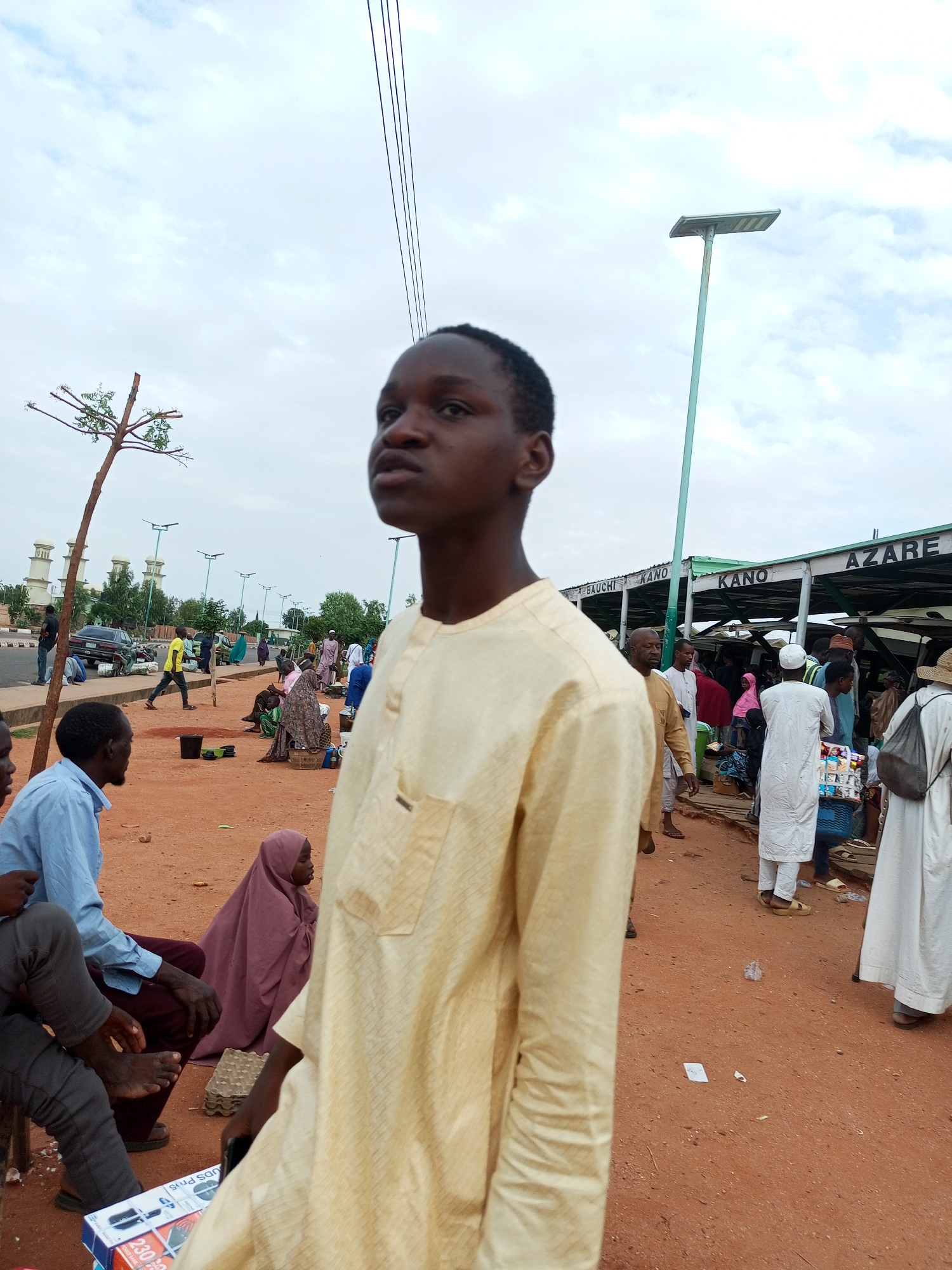 The image depicts a busy outdoor market scene in the city of Gombe, Nigeria, likely a motor park or a commercial hub. In the foreground, a young man in a pale yellow traditional garment is prominently featured, looking slightly upwards and to his left, with his face in profile. Behind him, the ground is covered in reddish-brown earth.
Several other people are visible in the mid-ground and background, engaged in various activities. Some individuals are seated, while others appear to be standing or walking. There are men and women, many of whom are wearing traditional African clothing, including headscarves and long robes. Objects such as baskets, a bucket, and a box containing electronic devices are scattered on the ground.
Overhead, power lines are strung across the sky, and a solar-powered street lamp is visible on a tall pole. In the distance, buildings with distinctive architectural features, possibly a mosque or government structures, can be seen. A sign with text that appears to include "BAUCHI KANO" and "AZARE" is affixed to a covered structure, suggesting different destinations or areas within the market. A car and motorcycles are also present, indicating transportation is a significant part of the scene. The overall atmosphere appears to be one of an active and bustling marketplace under a cloudy sky.