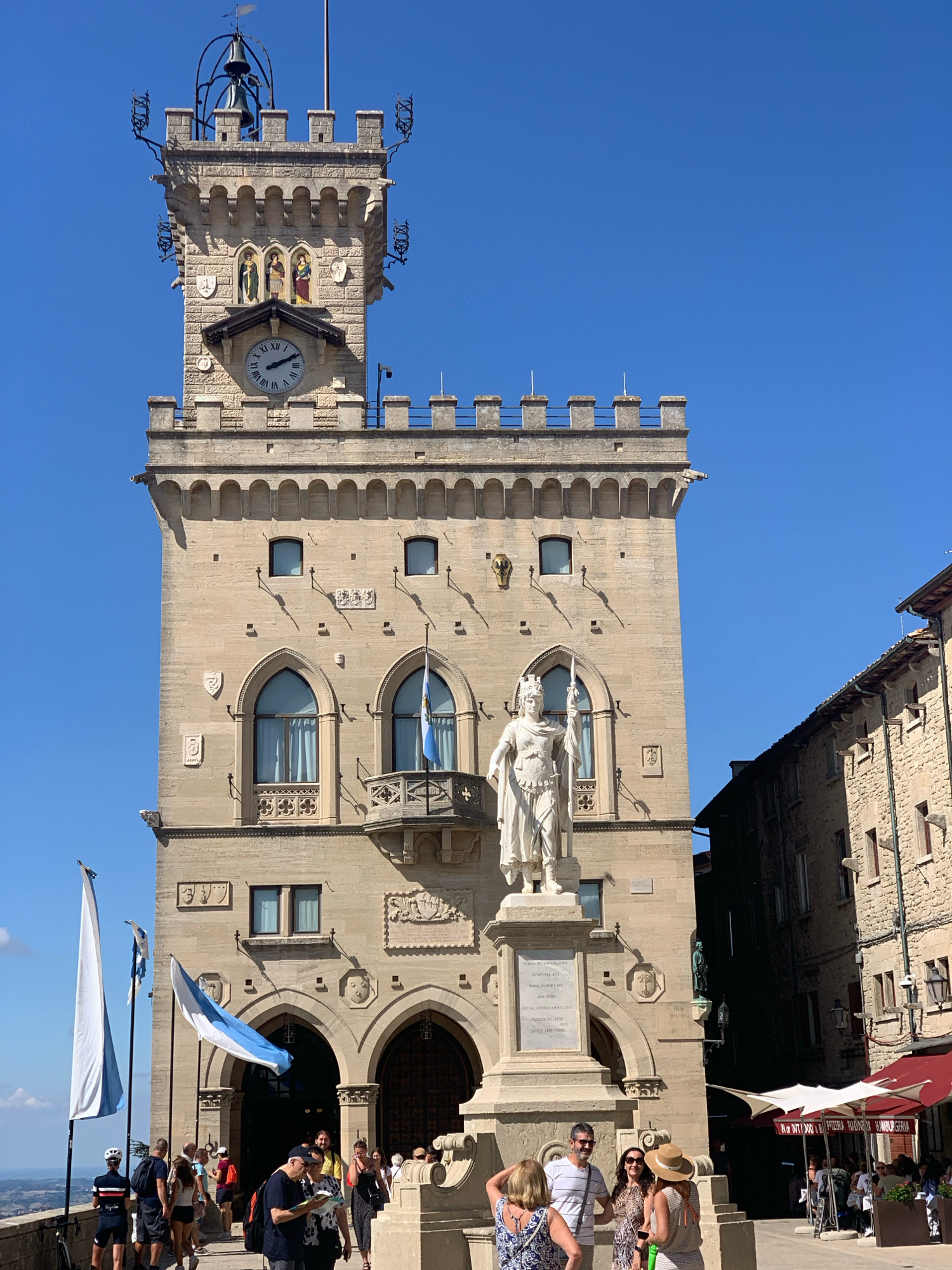 The image shows the Palazzo Pubblico in the City of San Marino, San Marino. The building is a tall, stone structure with a clock tower. A statue of Saint Marinus stands in front of the building. Several people are gathered in front of the building, and there are flags flying. The sky is clear and blue.