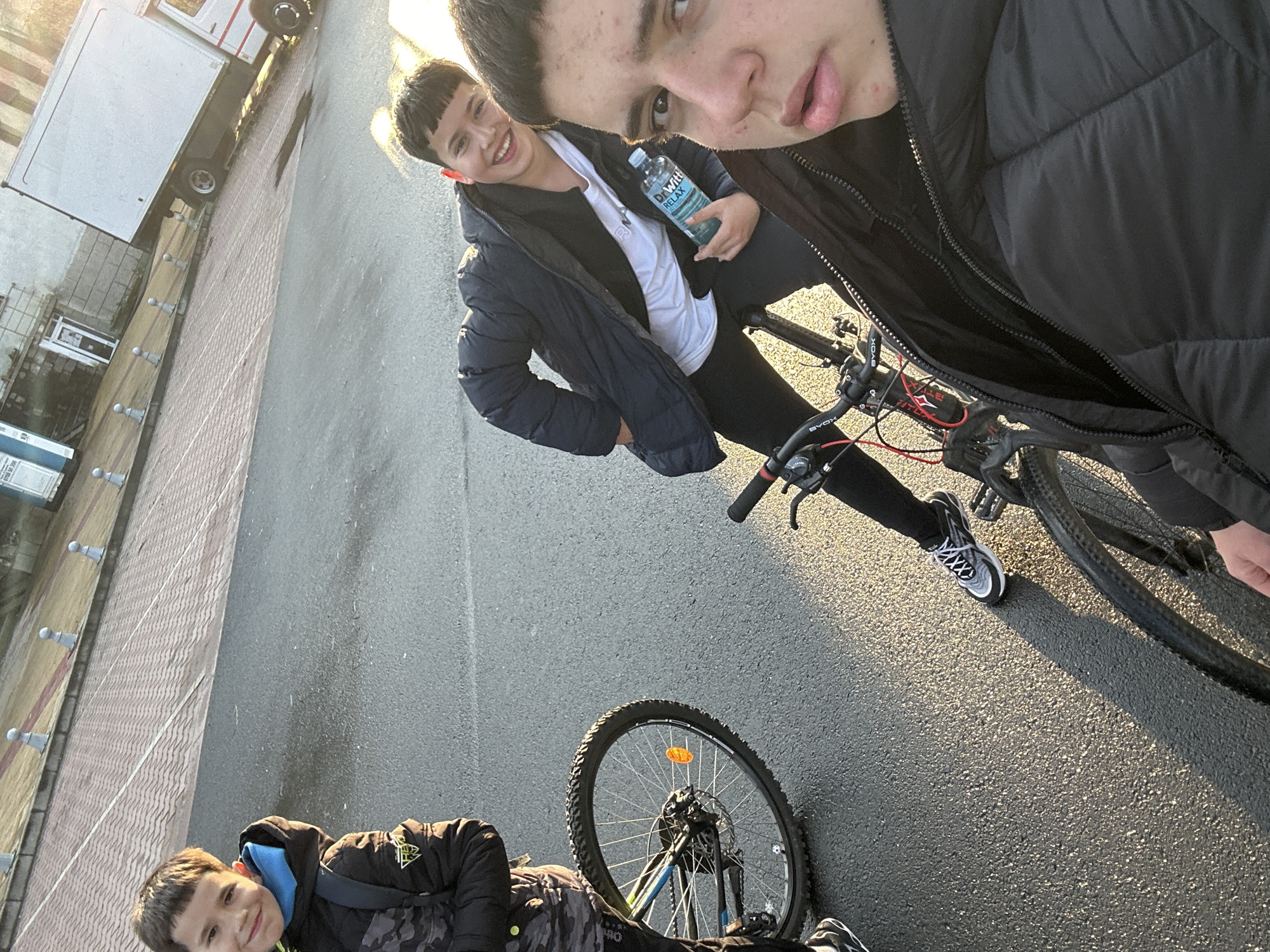 Three young people are on an asphalt road next to a brick sidewalk, possibly in Zlatograd, Bulgaria. Two are standing next to bicycles, one holding a water bottle, and the third is lying on the ground next to a bicycle. They all appear to be taking a selfie. A white truck is parked in the background. The sun is shining, casting a warm light on the scene.