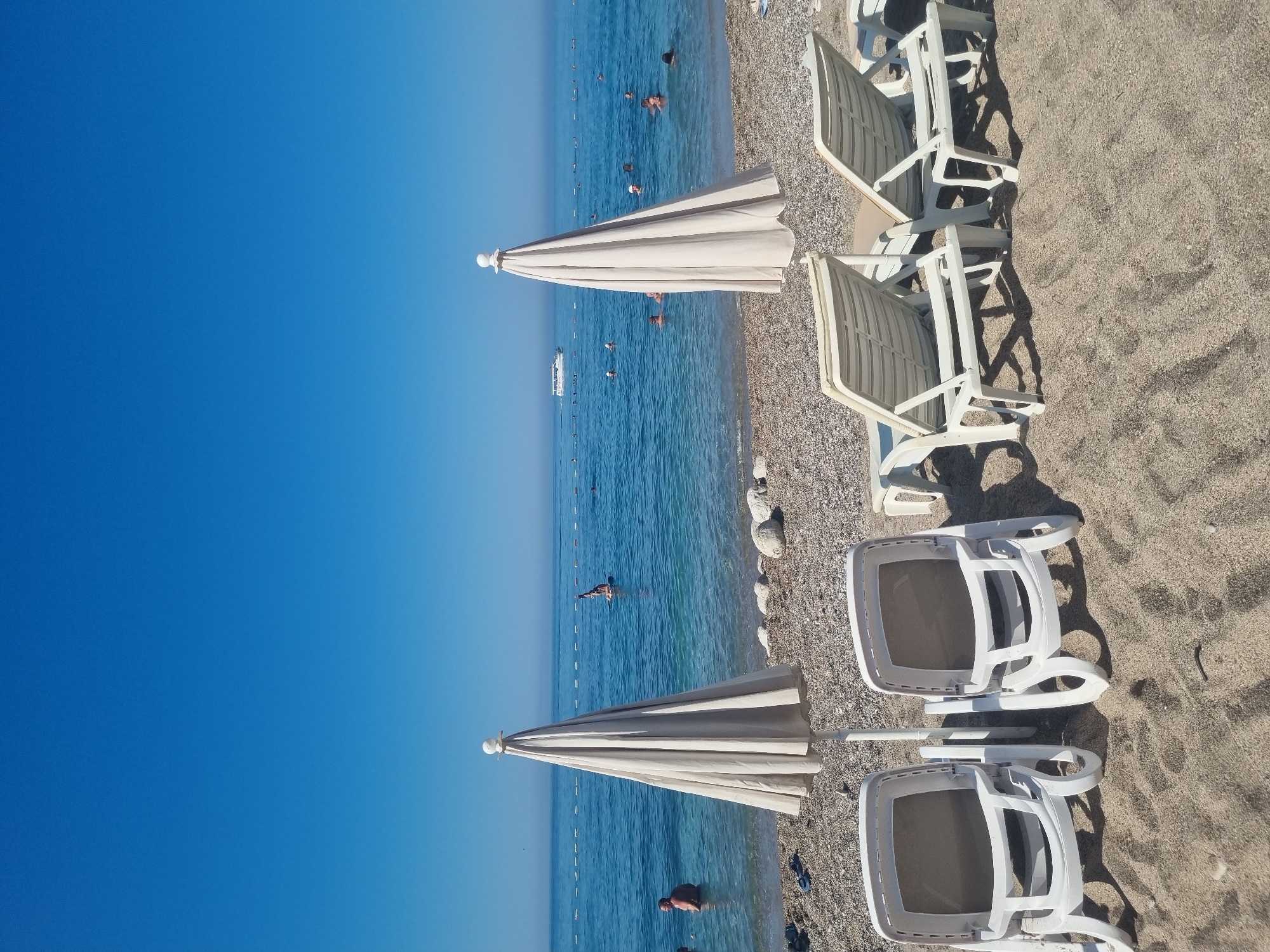 The image depicts a beach scene on a clear, sunny day in Sutomore, Montenegro. In the foreground, two closed, light-colored beach umbrellas stand on a pebble and sand shore. In front of the umbrellas are four white plastic lounge chairs arranged in pairs, facing the sea. The chairs are empty.
The sea is a vibrant blue, with gentle waves. Several people are visible in the water, some swimming and others standing. A person is seen on a paddleboard further out in the sea. A white boat is traveling on the horizon. A rope barrier with buoys stretches across the water, marking a section of the beach. The sky is a clear, cloudless blue. The overall atmosphere is one of a tranquil summer day at the beach.