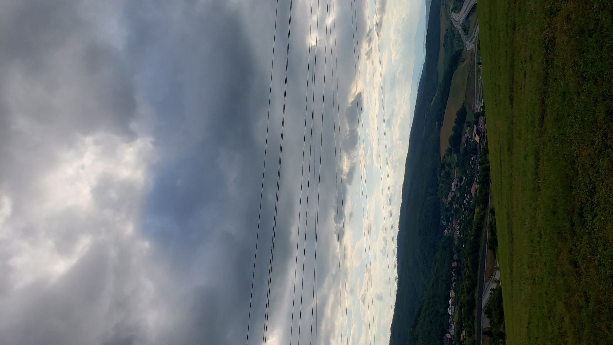 This image captures a landscape scene, likely in Prešov, Slovakia. The viewpoint is from a grassy hillside, looking towards a valley. Dominating the left side of the frame are dark, heavy clouds, suggesting overcast or approaching stormy weather. Several power lines run vertically through the middle of the image, stretching across the sky.
To the right of the power lines, the sky transitions to lighter, partly cloudy conditions with patches of blue sky visible. The valley below is densely forested on one side, with a small town or village nestled amongst the trees. Houses with red and brown roofs are visible. A highway curves through the valley, with cars on it. The right side of the image is a brightly lit, green, grassy field, likely the hillside from which the photo was taken.
The time of day is difficult to pinpoint precisely due to the cloudy conditions, but the lighting suggests it could be late afternoon or early evening. The overall mood is somewhat dramatic due to the stark contrast between the dark clouds and the bright, green landscape. There is no visible text in the image.