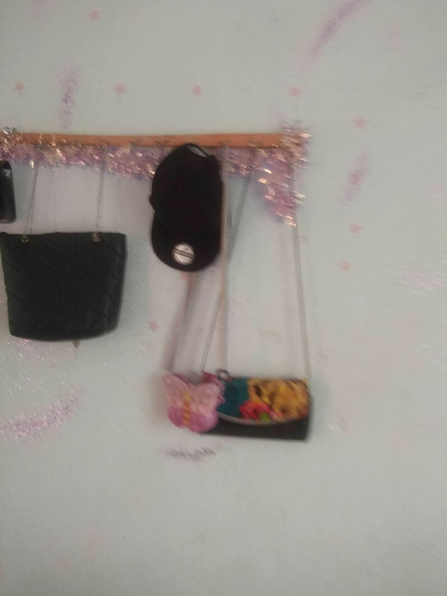 The image shows a collection of handbags hanging on a wooden rack against a light blue wall. The rack is decorated with pink tinsel. The handbags include a black quilted purse, a black cap, and a small clutch with a butterfly embellishment and a floral pattern. The background wall has faint pink star-shaped markings and some smudged pink glitter. The scene appears to be an indoor display, possibly in a closet or a personal space, within Jalingo, Nigeria. There are no people visible in the image.
