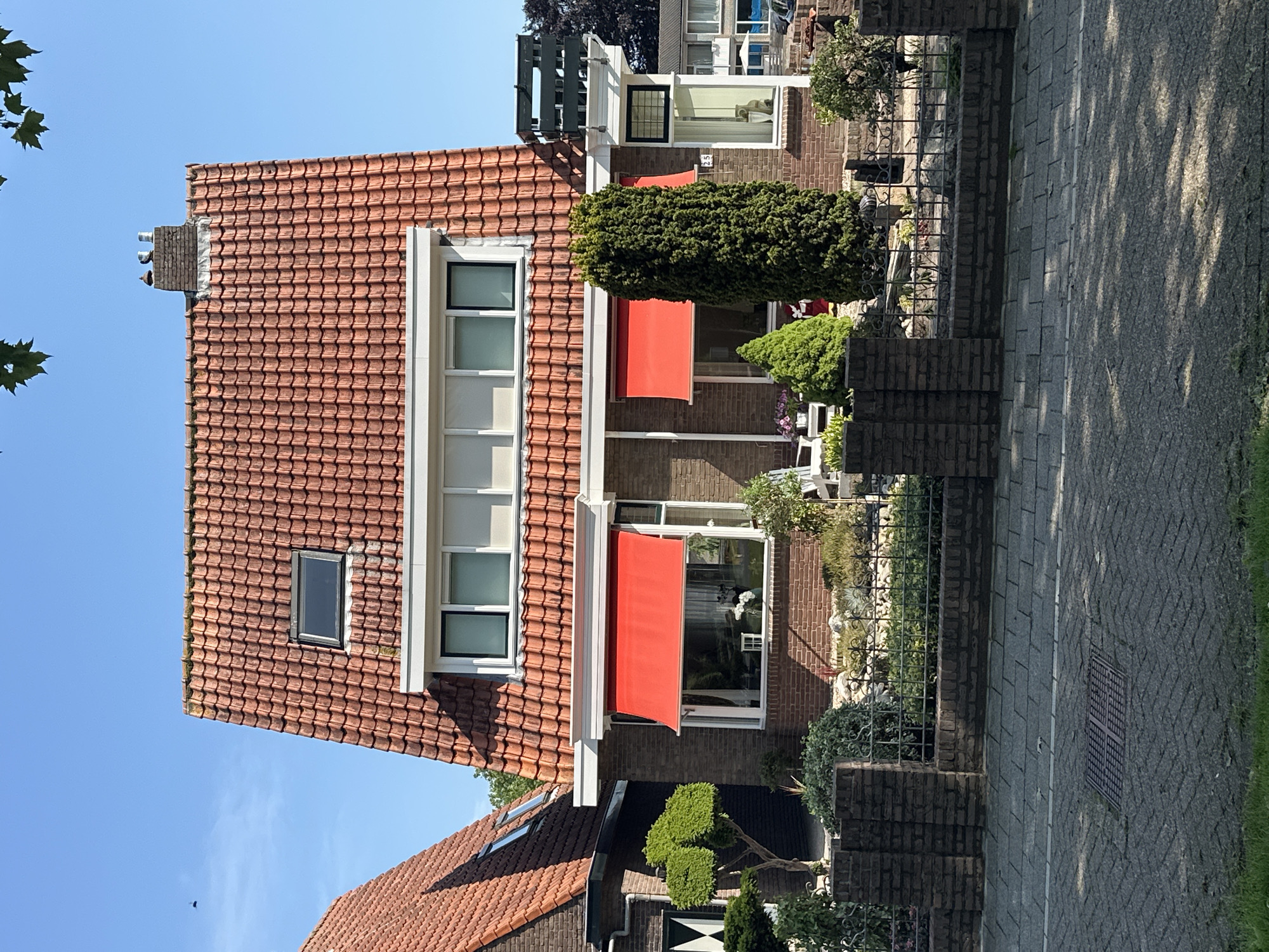 The image shows a detached brick house with a red tiled roof, located in Steenwijk, Netherlands. The house features multiple windows, some with red awnings, and a dormer window on the roof. A chimney with two objects on top is visible. The property is surrounded by a low brick wall and a decorative metal fence. The garden area contains various plants, shrubs, and a sculpted tree. In the foreground, a paved sidewalk with a drain cover is visible, and the street is lined with trees casting shadows. The sky is clear and blue. There are no people visible in the image.