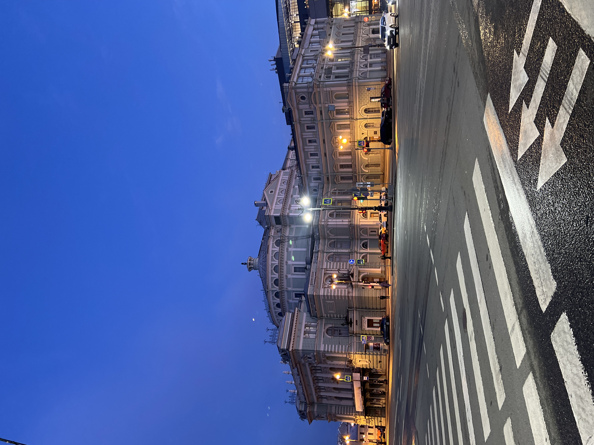 The image depicts a street scene in Saint Petersburg, Russia, at dusk or dawn, characterized by a deep blue sky. The Mariinsky Theatre is prominently featured on the left side of the frame, its architectural details illuminated by streetlights. A crosswalk with white stripes and arrow markings dominates the foreground, indicating pedestrian access across the wet, reflective asphalt road. Streetlights cast a warm glow along the edge of the road, illuminating parked cars and pedestrian traffic. The overall scene conveys a serene urban setting.