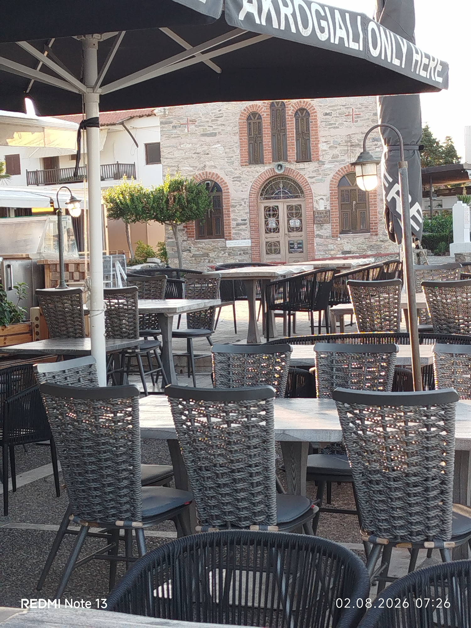 The image depicts an outdoor seating area of a restaurant or cafe, likely in Sithonia, Greece, given the architectural style and the presence of an umbrella with the inscription "AKROGIALI ONLY HERE." The foreground is dominated by several tables and chairs made of wicker and metal, arranged on a paved surface. Sunlight filters through the umbrellas and the woven backs of the chairs, creating a dappled light effect. In the background, a stone building with arched windows and doors, possibly a church or historical structure, is visible. The scene is serene, with no people present, suggesting it is either early morning or late afternoon. A street lamp with a lit bulb stands to the right of the central umbrella pole. The overall impression is one of a tranquil European outdoor dining space.