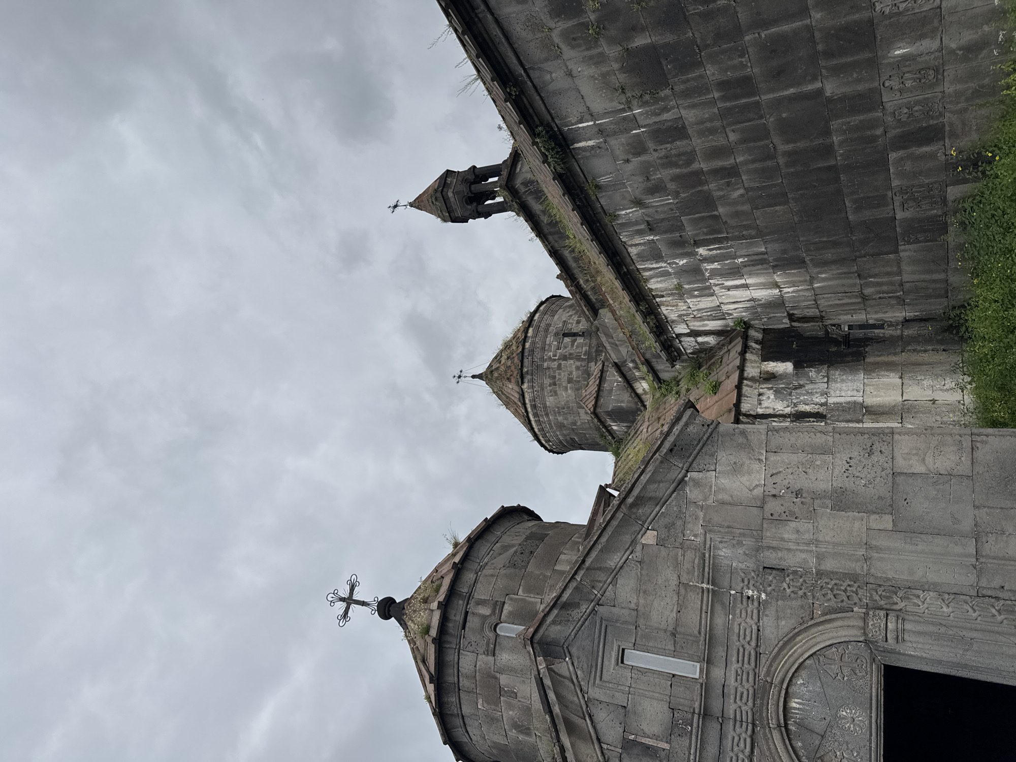 The image shows a low-angle view of the Haghpat Monastery in Haghpat, Armenia, under a cloudy sky. The ancient stone architecture of the monastery dominates the frame, with its weathered grey stone blocks and tiled roofs. Several domes and a bell tower are visible, each topped with a cross. Green vegetation grows on the roofs and along the walls of the structures, suggesting the passage of time and the natural encroachment of the environment. The details of the stone carvings around an arched entrance are visible on the left side of the image. The overall atmosphere is solemn and historic.