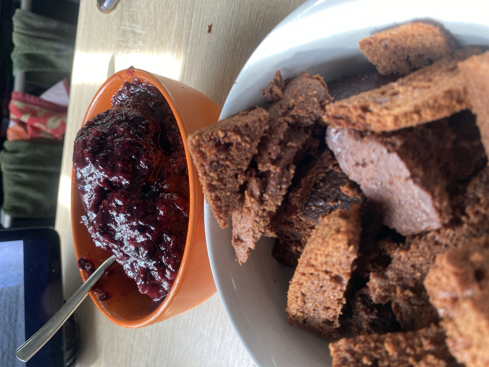 On a wooden surface, there's an orange bowl filled with dark berry jam or compote with a silver spoon. Next to it, a white bowl is brimming with chunks of brown cake or gingerbread. The scene appears to be indoors. A glimpse of what might be a tablet or phone screen is visible in the lower left corner of the frame. Details suggesting a specific location in Chaikovsky, Russia, are absent.