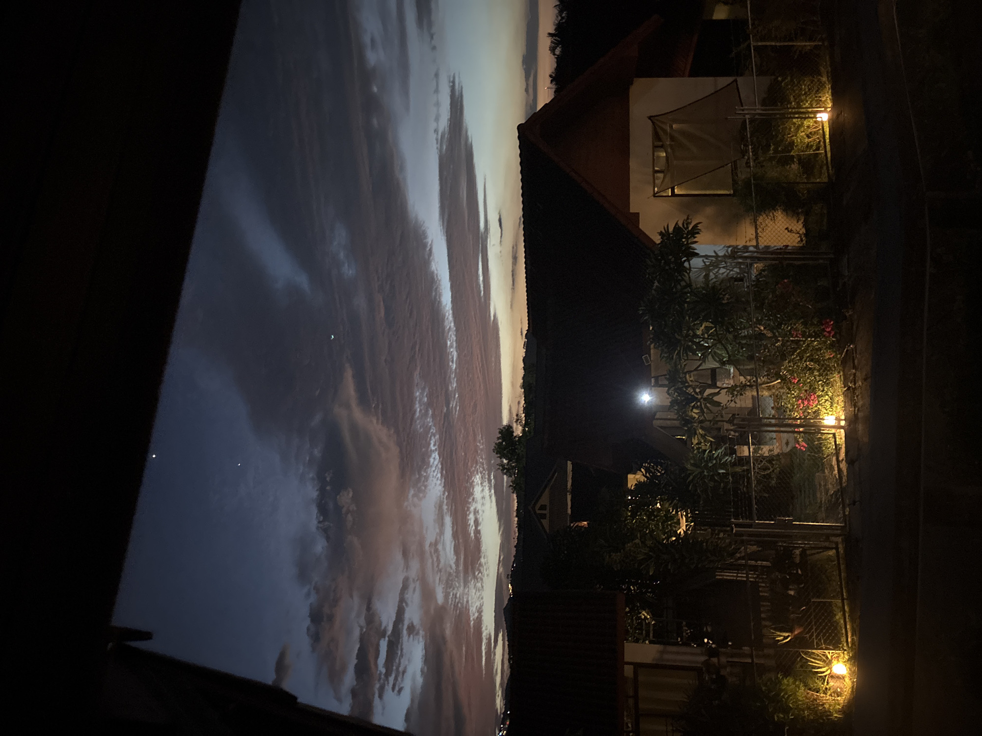 In the city of Baan Bang Rak, Thailand, the image captures a twilight scene from a high vantage point, looking out over a residential area. The sky is a gradient of deep blues and purples, with scattered clouds tinged with pink and orange from the setting sun. Two bright celestial bodies, likely planets or bright stars, are visible in the upper portion of the sky, along with a sliver of a crescent moon. Below the sky, the dark silhouettes of rooftops of several houses are prominent, with the distinct gabled roof of one house centrally positioned. Trees and foliage are visible in the foreground and between the houses, some illuminated by the warm glow of outdoor lighting. Several small, bright yellow outdoor lights are placed along a fence line in the lower section of the image, illuminating plants and a pathway. The overall impression is one of a peaceful evening in a tropical residential setting.