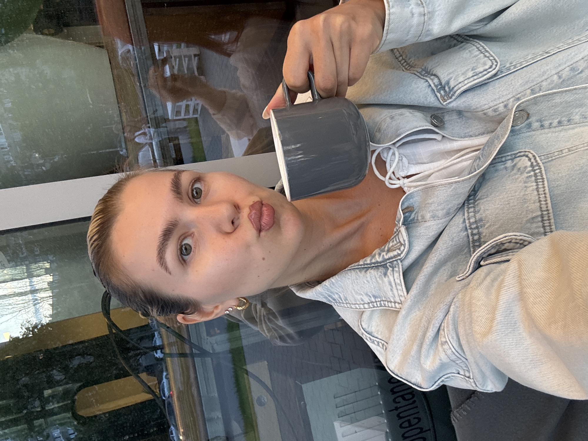 In an indoor setting with a large window, a young woman is captured in a selfie. She is holding a grey mug in her right hand and has her lips pursed. She is wearing a light blue denim jacket over a white top. The window behind her reflects the outside environment, showing cars on a street and buildings. The lighting suggests it is daytime.