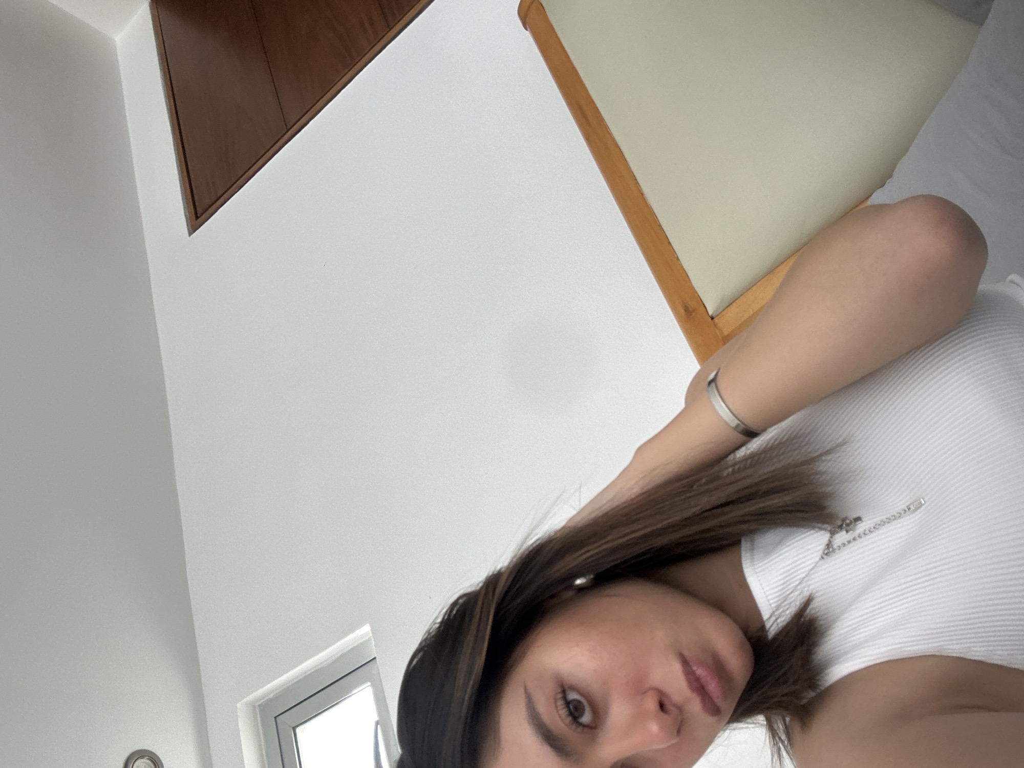 The media file displays a close-up, angled indoor shot, likely a selfie. The primary subject is a young woman, partially visible from the chest up, looking directly at the camera. She has long, dark brown hair that falls across her shoulder and chest. She is wearing a white ribbed sleeveless or short-sleeved top, and a silver bracelet is visible on her left wrist. A delicate silver chain necklace with a small, possibly safety-pin-shaped pendant, is also visible on her top. Her expression is neutral to slightly serious, with dark eyebrows and eyes.
The background consists mainly of white walls, suggesting a bright room. In the upper left corner, there's a dark brown wooden structure, possibly a wall panel, ceiling feature, or furniture. Another wooden element, lighter in tone, and part of a cream-colored upholstered chair are visible in the upper right. A window frame with light visible through it is in the lower-left corner of the image, indicating it is daytime. Below the window, a small circular metallic object, possibly a light fixture or part of a clock, is partially visible. No discernible activity beyond posing for the photograph is evident. There is no visible text in the image.