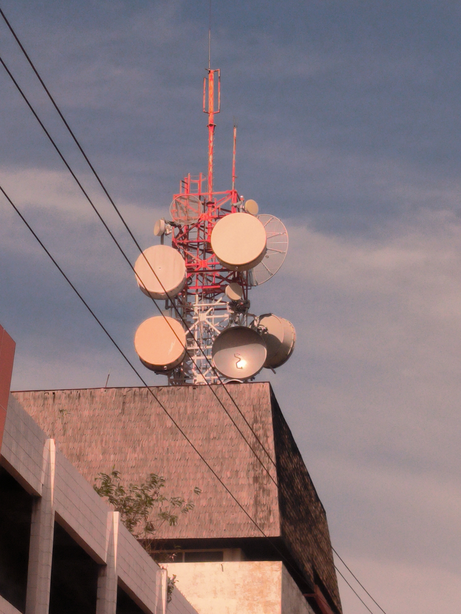 A tall, red and white telecommunications tower stands atop a building in Lhokseumawe, Indonesia. The tower is equipped with multiple circular microwave antennas of various sizes. Overhead electrical wires pass in front of the tower. The building below appears weathered, with a pattern resembling tiles on its upper portion. Adjacent is a newer-looking building with white architectural features. A small tree grows near the base of the tower, and the sky is overcast.