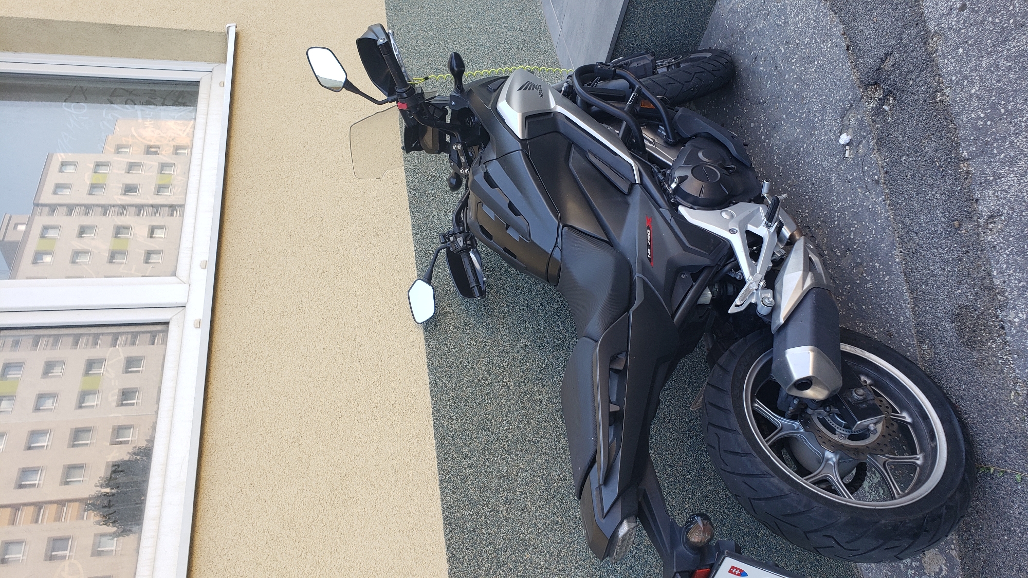 A black Honda NC750X motorcycle is parked on a concrete surface in front of a building. The building has a textured yellow facade on the upper portion and a dark grey speckled facade on the lower portion, separated by a white horizontal trim. A large window is visible on the upper part of the building, reflecting an apartment building. The motorcycle is positioned diagonally, facing away from the viewer and slightly to the right. It has a black seat, black bodywork, silver accents, and a silver exhaust pipe. The rear tire is large and black, with a silver rim. A Slovakian license plate is visible on the rear of the motorcycle. The ground is a rough, grey concrete with some debris. The scene appears to be an outdoor urban environment.