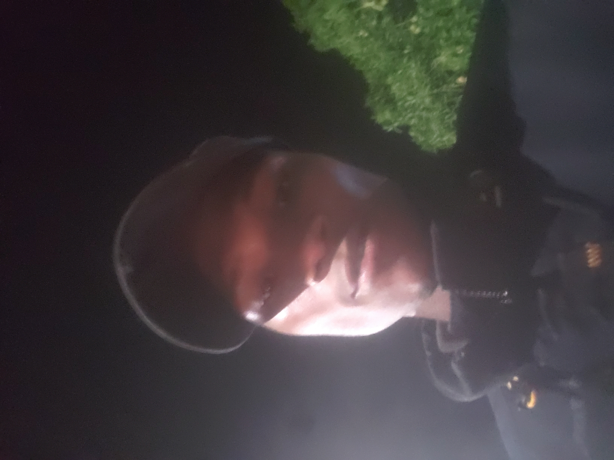 A dimly lit, medium-range shot depicts a person wearing what appears to be a police or security uniform and a cap, likely outdoors. The setting is dark, but some greenery is visible in the background. Details of the uniform are unclear due to poor lighting. The location, Bukuru, Nigeria, is not confirmable from the image alone.