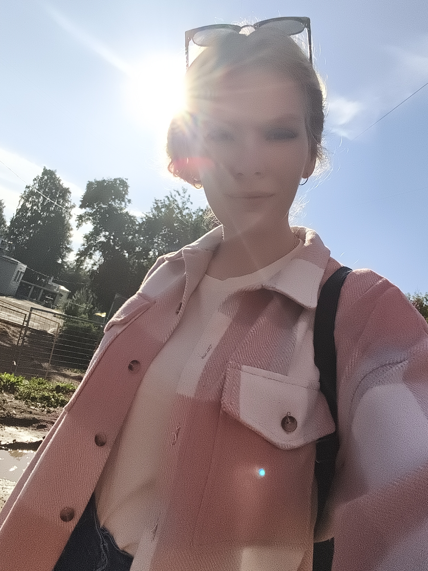 A young woman is captured in a selfie-style photograph, bathed in bright sunlight. She has blonde hair pulled up, with sunglasses perched on top. She's wearing a pink and white plaid shacket over a light-colored top. A black strap, likely from a backpack, is visible over her shoulder. Her expression is neutral.
The background suggests an outdoor setting, possibly a backyard or rural area. There are trees in the distance, and a fence along a dirt path with some muddy puddles. A small building or shed is visible to the left, and a power line stretches across the blue sky. The strong sunlight indicates it is daytime, likely a bright and clear day. The presence of puddles could suggest recent rain.
There is no visible text in the image. The overall mood is casual and bright, capturing a moment in an outdoor environment.