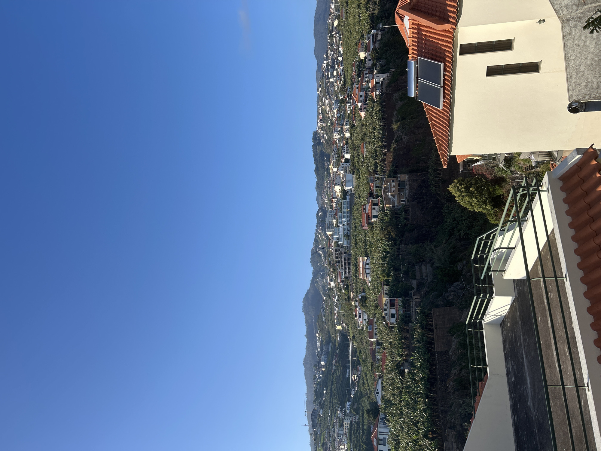 The image shows a sunny landscape in Câmara de Lobos, Portugal. The sky is clear and blue. A hillside densely populated with buildings and vegetation dominates the center of the frame. The architecture appears to be mostly residential, with white buildings and terracotta rooftops. In the foreground is part of a building with a reddish-tiled roof, solar panels, light yellow walls, and a balcony with metal railings.