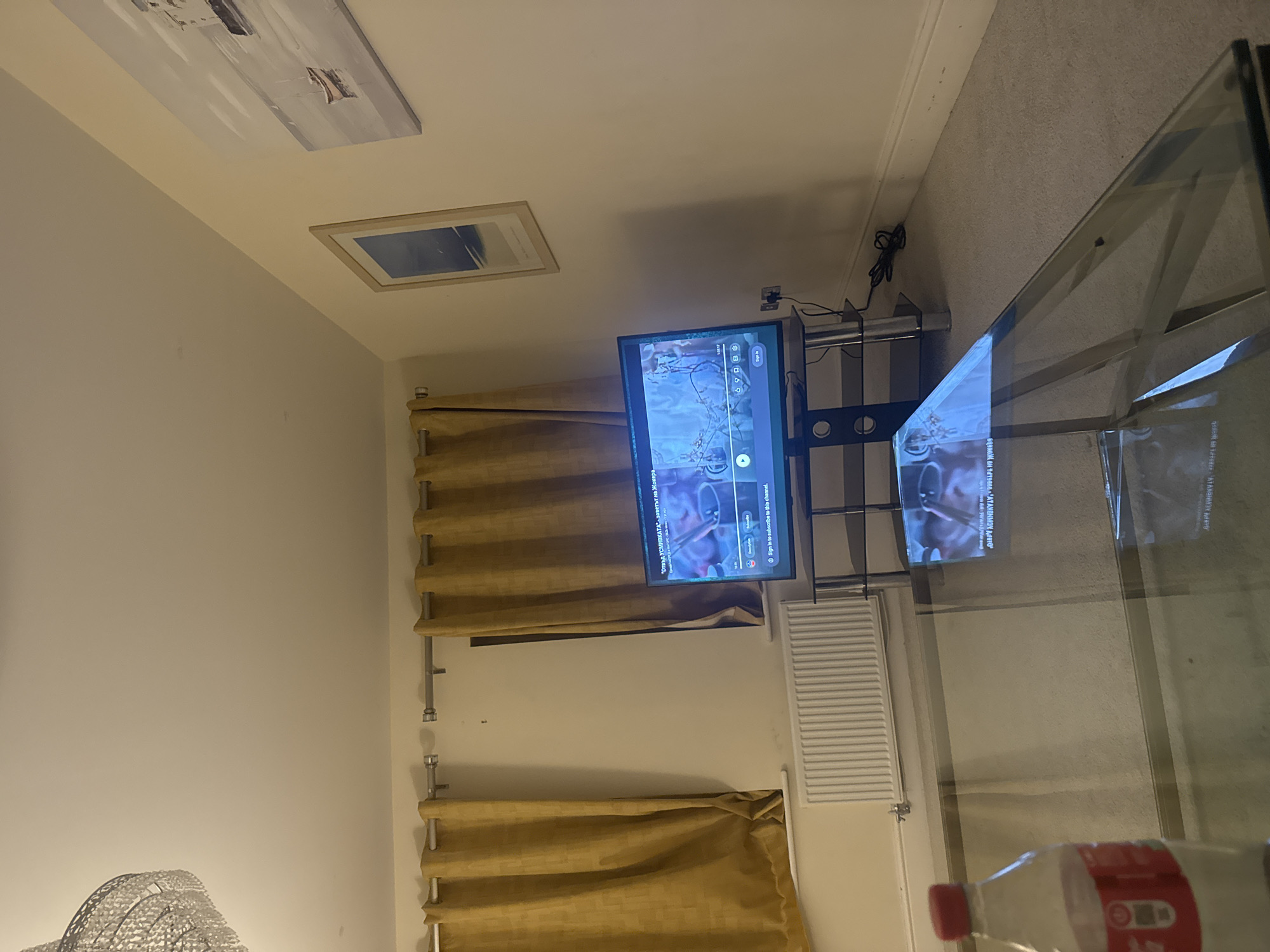 The image shows an interior scene. In the foreground is a glass coffee table. A bottle of water is partially visible on the table. A television is positioned on a stand against a wall, displaying what appears to be a video. Behind the TV, curtains hang on either side of a wall-mounted radiator. Two framed pictures hang high on the wall. The image is rotated counter-clockwise.