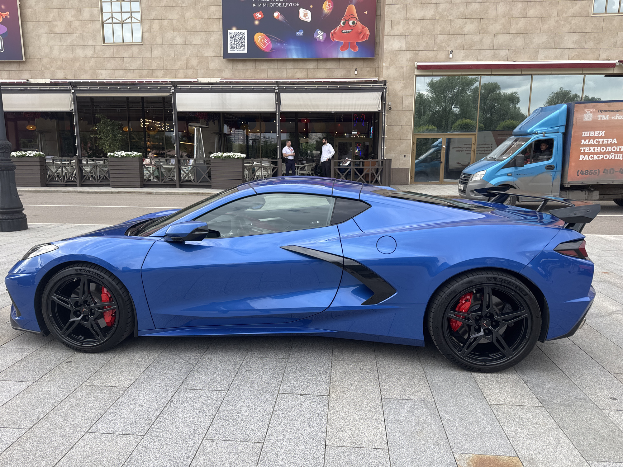 A bright blue Chevrolet Corvette C8 sports car is parked on a stone-tiled sidewalk in front of a building with a large billboard. The building has large windows, some of which appear to be part of an outdoor seating area for a cafe or restaurant. A blue and grey delivery truck is parked to the right of the Corvette. There are several people visible inside the restaurant area and one person in the delivery truck. The billboard above the building displays colorful graphics and text in Russian. The overall scene suggests an urban environment in Moscow, Russia, likely a commercial or entertainment district.