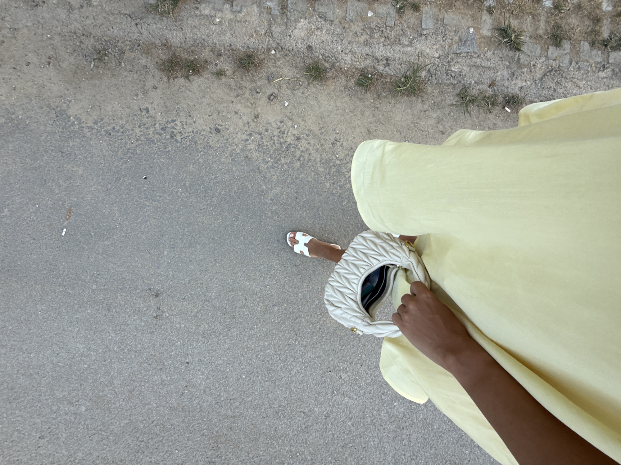 A person's hand, arm, and leg are visible from a high-angle, overhead perspective in this image, taken on a sidewalk in Ofrynio Beach, Greece. The person is wearing a pale yellow dress and white sandals. They are holding a cream-colored, pleated handbag with a braided strap. The ground is a textured asphalt surface with scattered debris. The overall scene is casual and outdoor.