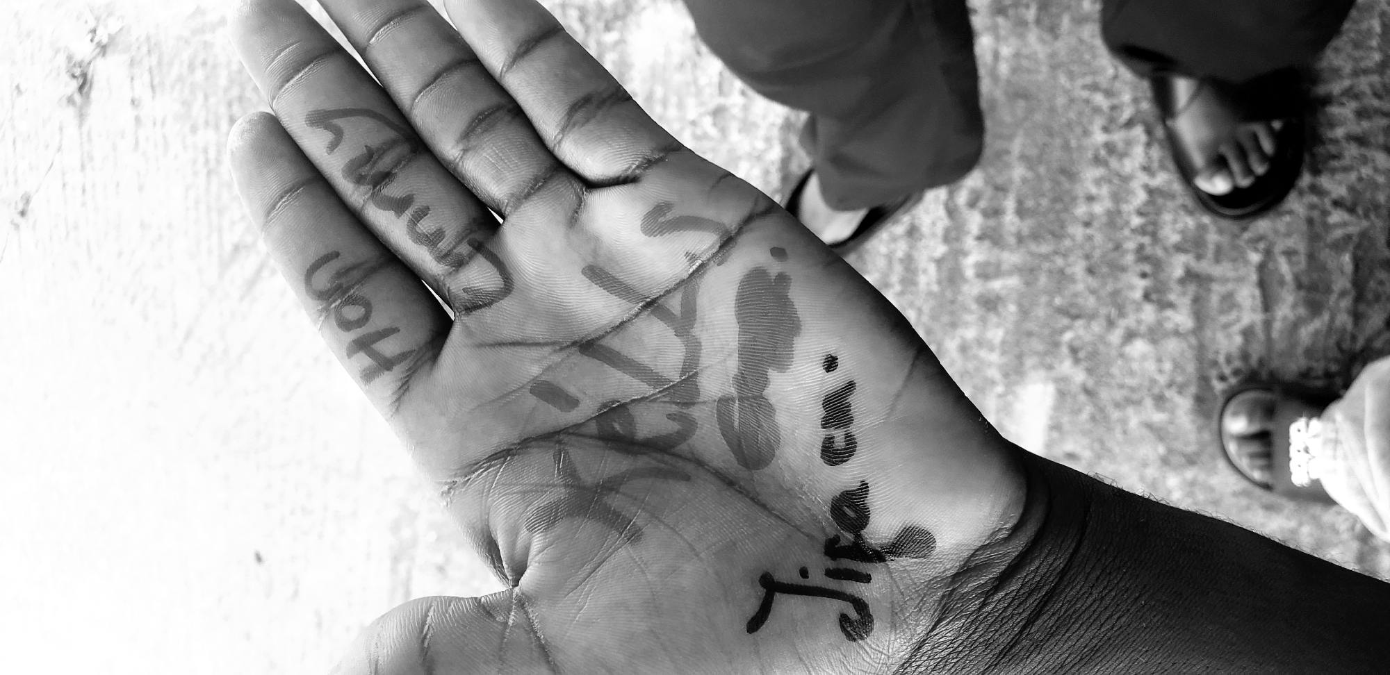 A black and white, close-up, eye-level shot shows a person's open palm, on which text is written in black marker. The text on the palm reads "Jija cm". Further up the fingers of the same hand, other text is visible but partially obscured and smudged. The finger text appears to say "Hon" on one finger and "ummy" on another. The hand is in focus, with the background out of focus. The background consists of a textured, concrete-like surface. Two sets of feet wearing sandals are visible at the bottom of the frame, suggesting the perspective of someone looking down at their hand. This image was taken in Barden Yana, Nigeria.
