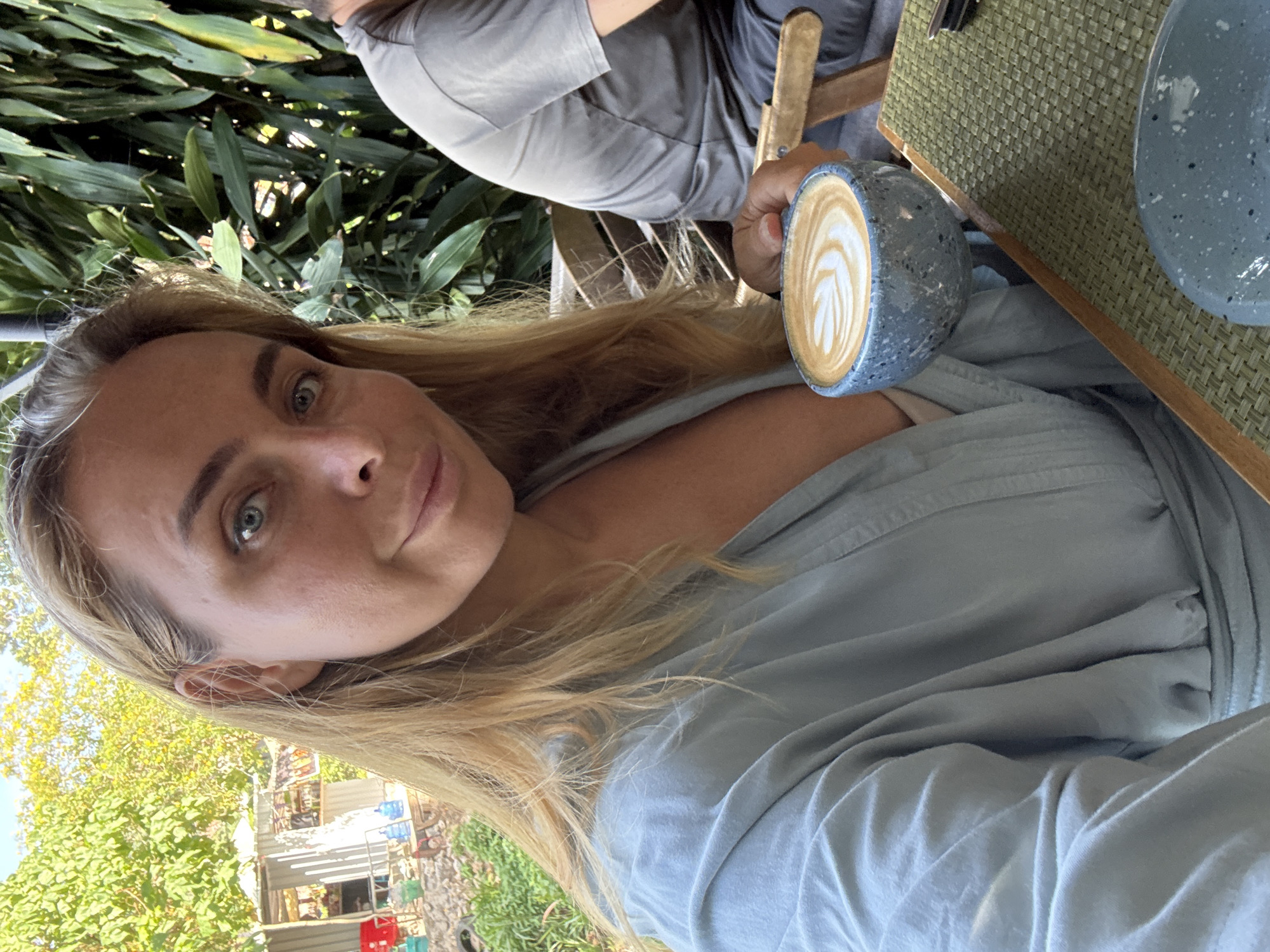 A close-up, selfie-style shot captures a blonde woman in Ko Samui District, Thailand, holding a blue speckled mug filled with latte art. She wears a light blue wrap or robe. Her eyes are open and she looks directly at the camera with a slight smile. To her right, a person in a gray t-shirt is partially visible, seated at a table with a woven green placemat. The background shows lush green foliage and what appears to be a small building with a red chair, suggesting an outdoor cafe or restaurant setting. The lighting indicates it is daytime.