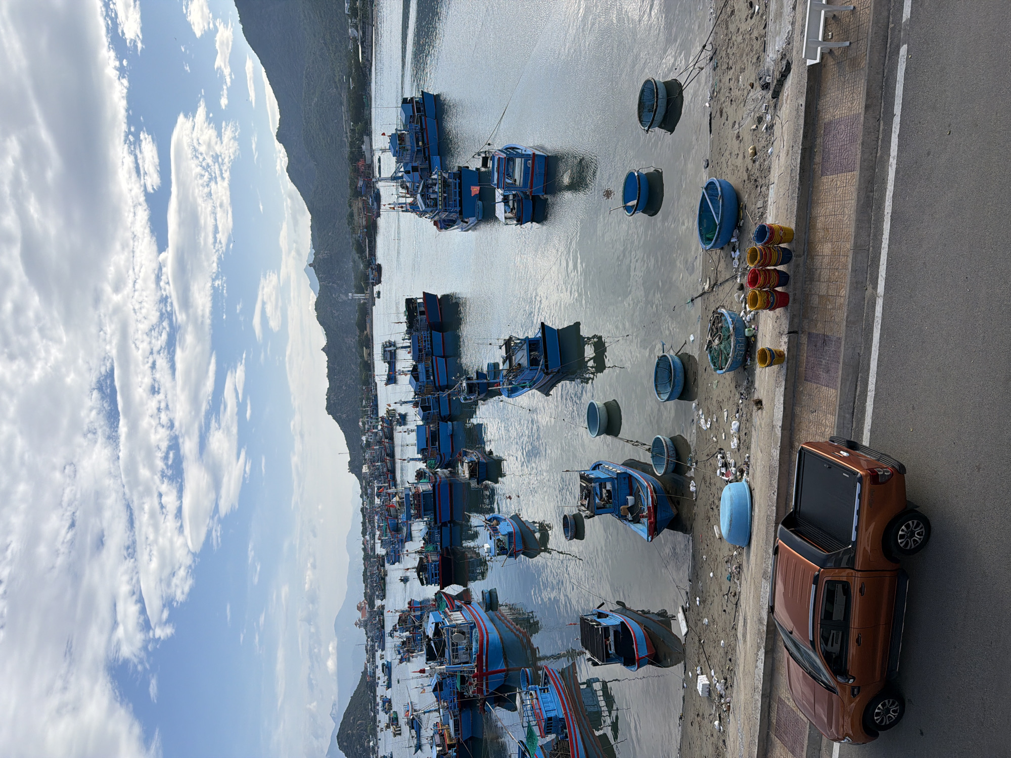 This image captures a vibrant fishing harbor or estuary in Ninh Chu Ward, Vietnam, under a bright, partly cloudy sky. The scene is dominated by numerous traditional blue wooden fishing boats, many with red trim, moored closely together in the calm, reflective water. Several iconic round basket boats, known as "thung chai," also dot the water and the muddy shoreline, some blue and some dark.
A paved road runs parallel to the waterway on the right side of the frame, separated by a concrete sidewalk and a muddy bank. An orange-brown Ford Ranger pickup truck with a dark tonneau cover is parked on this road. The bank itself is cluttered with various objects, including stacks of colorful red, yellow, and blue buckets or containers, as well as some scattered litter like plastic bottles and bags.
In the background, across the water, a range of verdant, hazy mountains stretches along the horizon, hinting at a coastal or riverine landscape. The sky is a clear blue adorned with large, fluffy white clouds, suggesting good weather conditions, likely late morning or early afternoon given the bright light.
The image portrays a typical bustling fishing community, where the boats are either awaiting their next journey or have recently returned from the sea. No specific human activity is visible, but the abundance of fishing vessels and gear points to an active local fishing industry. The primary identifiable text is the "Ford Ranger" branding on the pickup truck.