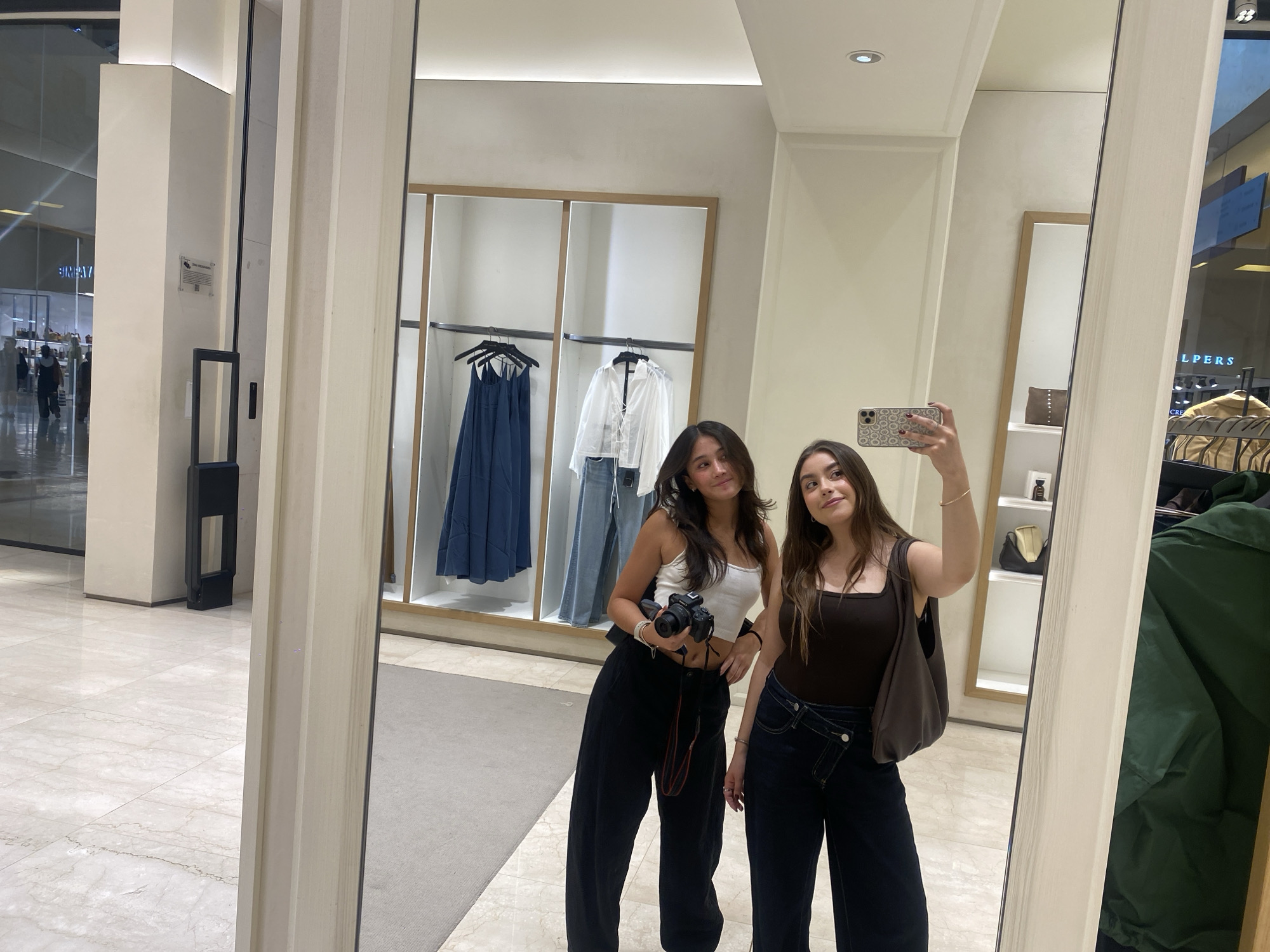 Two young women are reflected in a full-length mirror inside a retail store. The woman on the left, with dark hair, wears a white tank top and black pants, holding a camera. The woman on the right has dark hair and is wearing a brown tank top and dark jeans, holding a phone up to take a picture. Behind them, within the store, are clothing racks displaying various garments. Outside the store, visible through the entrance, is a brightly lit shopping area with other stores and people. The mirror's frame is off-white, and the store’s interior has a modern, clean design.