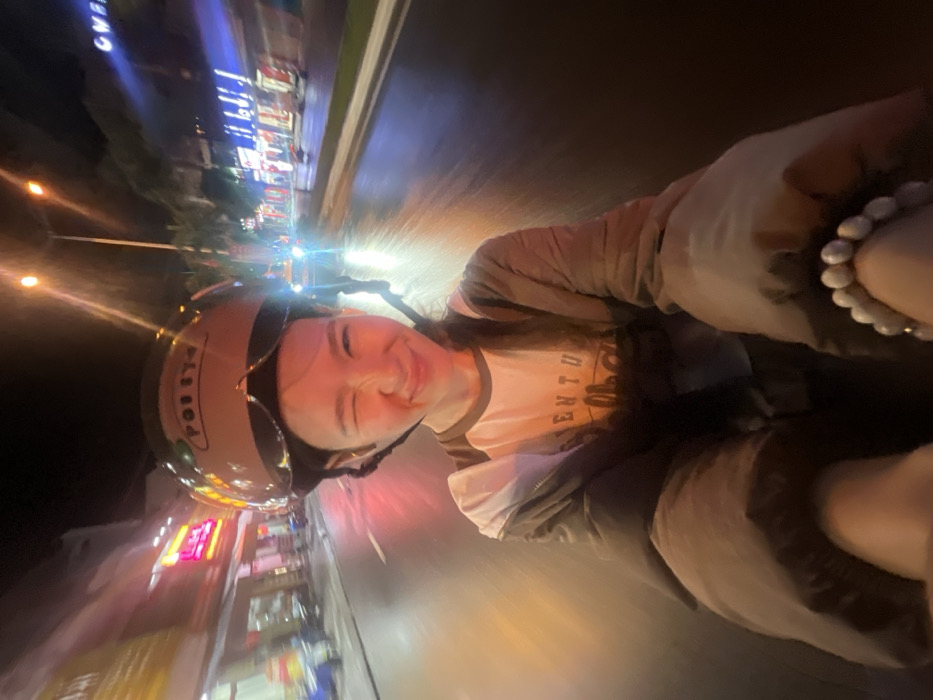 A self-portrait taken at night depicts a young individual riding as a passenger on a moving two-wheeled motorized vehicle on a wet urban street in Nha Trang, Vietnam. The subject, centered in the frame, wears a light brown helmet with a dark visor and an attached chin strap, a dark zippered jacket over a white and dark-sleeved raglan shirt with partial lettering "ENTU" visible, and a white beaded bracelet on her left wrist. She is looking directly at the camera, winking her right eye, and smiling. Motion blur affects the background, indicating movement. The multi-lane asphalt road is wet and reflects numerous artificial lights. On the left, blurry commercial storefronts display illuminated signs, including a prominent red sign with indistinct Vietnamese characters. On the right, additional commercial buildings feature blue and white lighting. Overhead streetlights cast an orange glow, and bright headlights from oncoming traffic are visible. The scene captures a bustling commercial thoroughfare in Nha Trang at night.