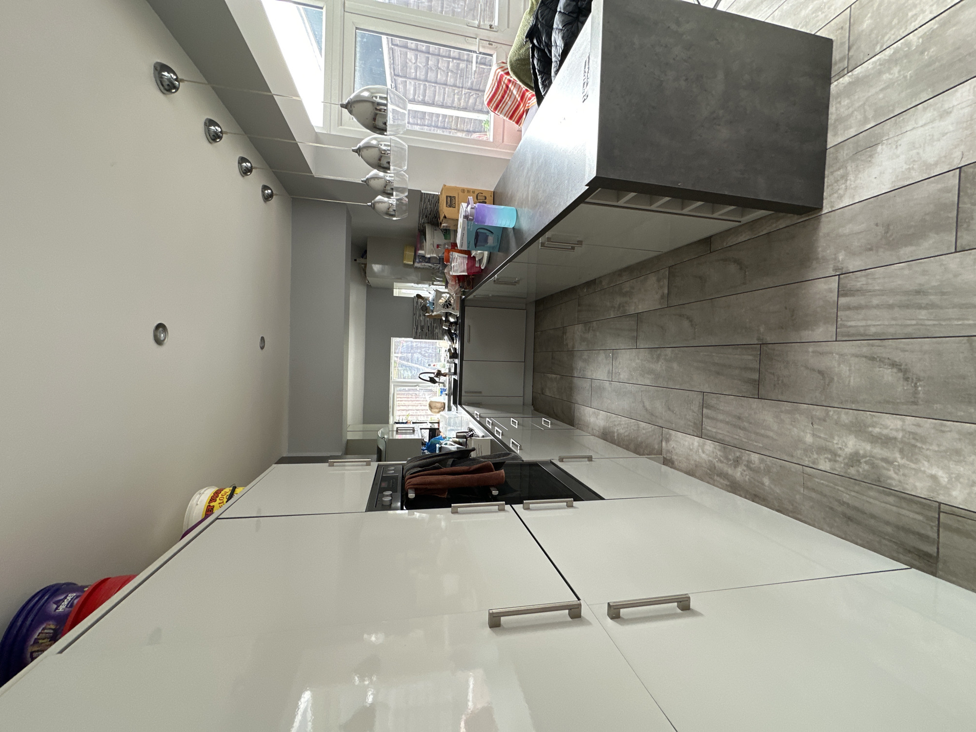 The image shows an interior view of a modern kitchen. Dominating the foreground are glossy white cabinets with silver handles. A dark gray countertop runs along the right side of the image. Above the countertop are light gray wood-look tiles. Towards the background, there's a window with natural light, offering a glimpse of the outside. Hanging from the ceiling are a series of pendant lights with a chrome finish. Other kitchen items like containers and utensils can be seen. There's no evidence to suggest this location is in London. No people or actions are apparent in the image.