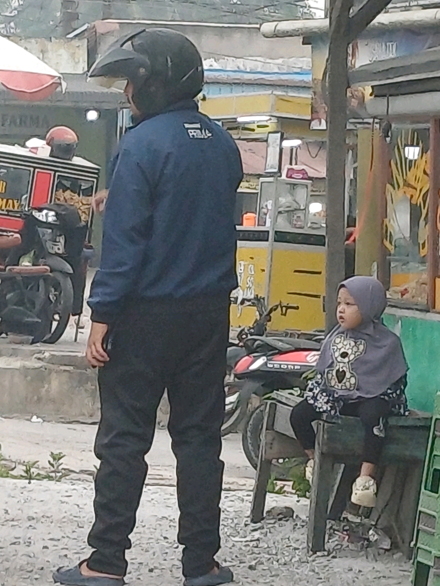 A man wearing a dark blue jacket, black pants, and a black helmet stands in the foreground, facing away from the camera. A young child wearing a grey hijab and a patterned shirt sits on a wooden bench to the right of the man. The child is looking to the left. In the background, a red and white food cart and motorcycles are visible. The scene appears to be an outdoor market or street vendor area, likely in Dumai City, Indonesia, given the presence of a food cart and motorcycles commonly seen in such settings. The ground is covered in gravel and there are some low-lying plants. The overall lighting suggests it is daytime.