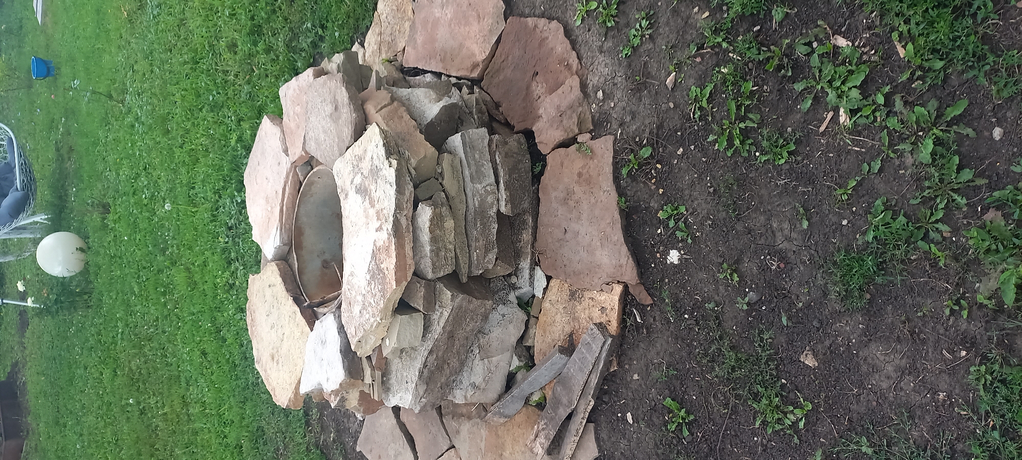 A fire pit constructed from stacked, irregularly shaped stones is visible in a grassy outdoor area, likely a backyard. The stones vary in color, ranging from light tan to reddish-brown, and are arranged to form a circular structure with a metal basin at its center. The ground around the fire pit is composed of dark soil with sparse green vegetation. In the background, out of focus, a white hammock hangs from a stand, a large white spherical object, and a blue bucket are present. The scene suggests a domestic outdoor setting.