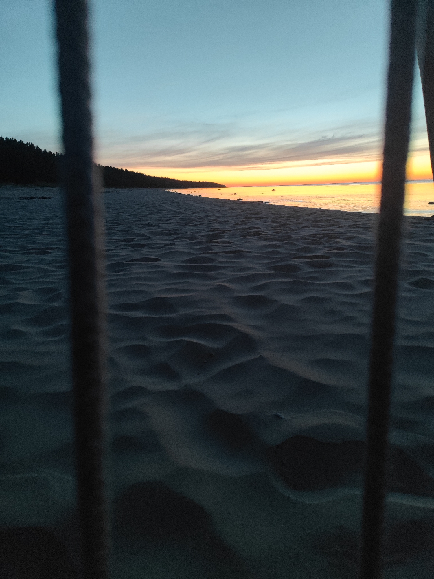 The image depicts a sandy beach in Motornoe, Russia, at dusk. The foreground is dominated by textured sand, showing ripples and undulations. Two vertical, dark, blurred objects, possibly posts or bars, obscure parts of the view. Beyond the sand, the calm sea stretches to the horizon, reflecting the warm hues of the setting sun. The sky transitions from a soft blue to vibrant oranges and yellows near the horizon, with subtle cloud streaks. A dark, forested shoreline is visible in the distance on the left. The scene is quiet and serene, capturing the transition from day to night.