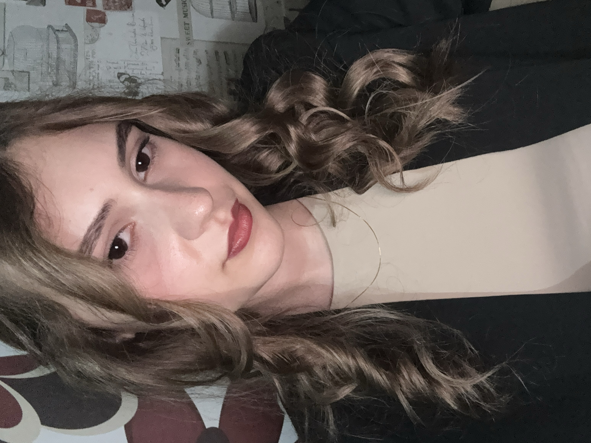 A close-up, slightly rotated selfie of a woman. She has fair skin, brown eyes, dark eyebrows, and wavy, light brown hair. She is wearing a black jacket over a beige top. A thin gold hoop earring is visible. The background consists of a patterned surface on one side and a wall with a repeating design reminiscent of vintage ephemera on the other. The location is not identifiable as the city of Panagyurishte, Bulgaria based solely on the provided image.