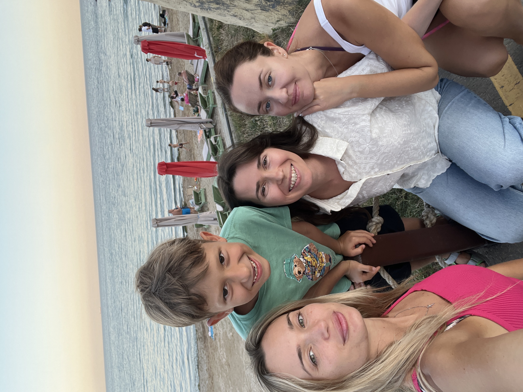 In Fontanka, Ukraine, a group of four people, two women and two children, are captured in a selfie on a beach. The younger woman on the left wears a vibrant pink tank top and a delicate necklace. Beside her, a boy with light brown hair smiles broadly, dressed in a green t-shirt featuring a teddy bear graphic. In the center, a woman with dark hair smiles gently, wearing a white, textured blouse and braces. To her right, another woman with light brown hair and blue eyes rests her head on her hand, her arm around the woman in the center, sporting a white tank top and a purple bikini strap visible. The background reveals a sandy beach with loungers and red umbrellas, leading to the calm blue sea under a soft, dusky sky.