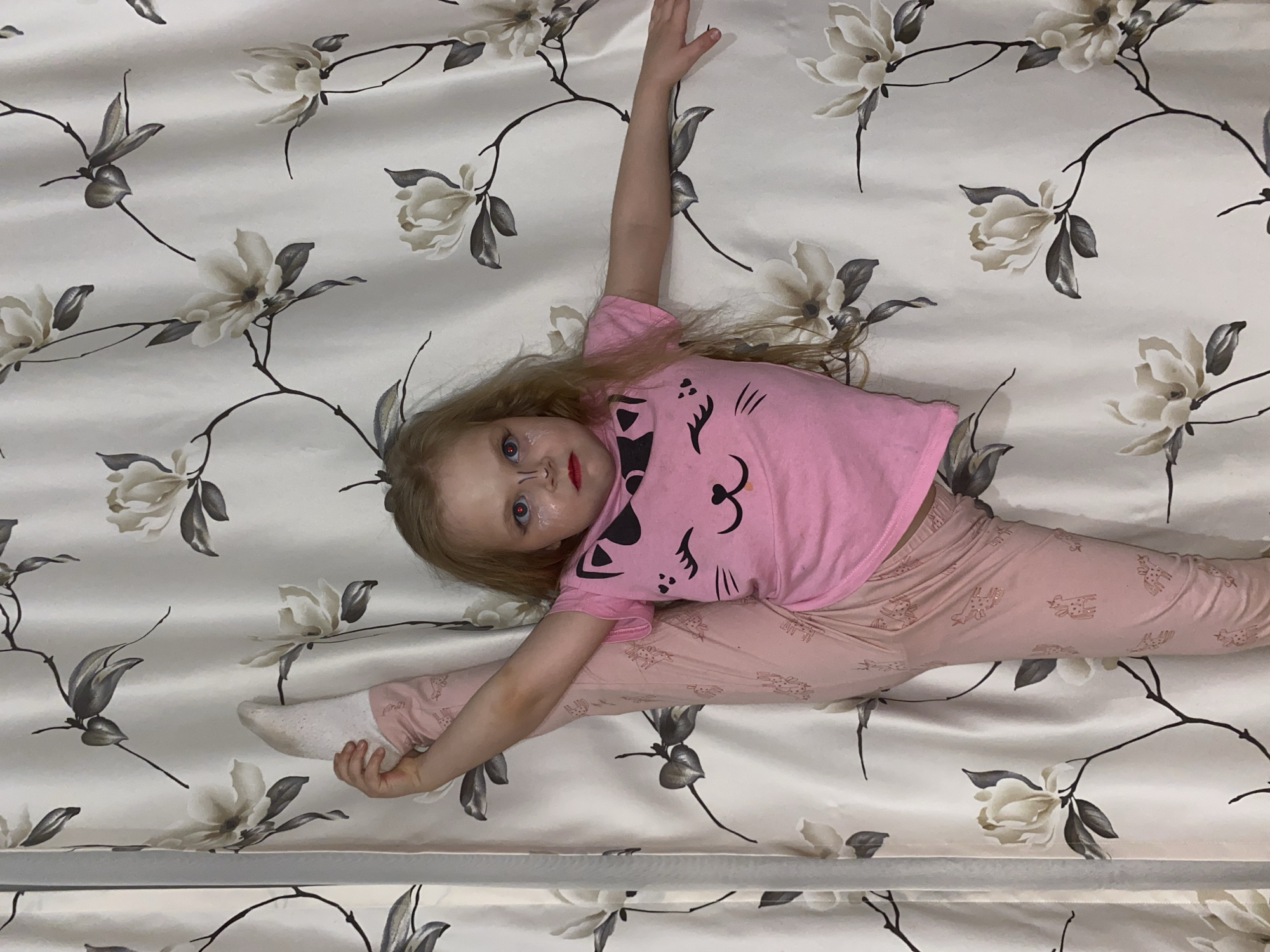 A young child lies on a bedspread patterned with cream-colored flowers and dark branches. The child is dressed in pink pajamas with a cartoon cat design on the shirt and giraffe patterns on the pants. They are performing a split, with one leg extended to the left and the other to the right. One foot is covered in a white sock, and the opposite hand is raised above their head. The child has blonde hair, blue eyes, and is wearing makeup, including drawn-on whiskers and red lipstick. The environment appears to be indoors. There is no indication of the city of Votkinsk, Russia.