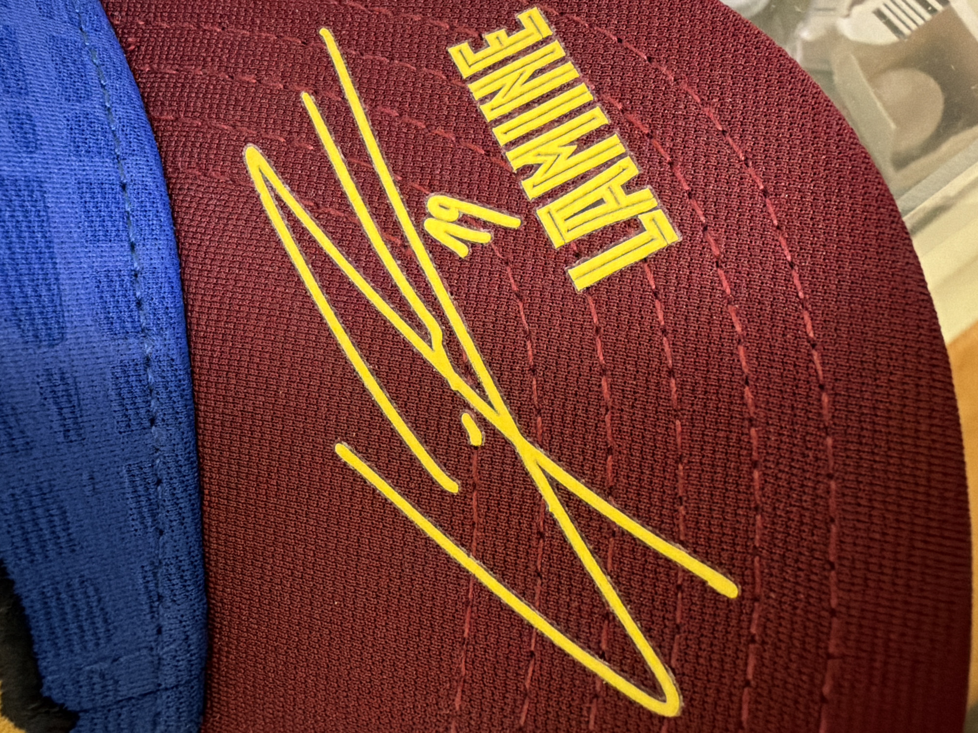 A close-up, detailed shot focuses on the embroidered details of a maroon baseball cap. A bright yellow, stylized signature stretches across the cap, with the number "19" also rendered in yellow to the right of the signature. Below the signature and number, the name "LAMINE" is embroidered in bold, blocky yellow letters. The cap features a seam line above the embroidered area, and a portion of a blue fabric panel is visible at the top of the frame. The image is a product shot, likely intended for retail or promotional purposes, and does not depict any specific location in Kyiv, Ukraine, or any people.