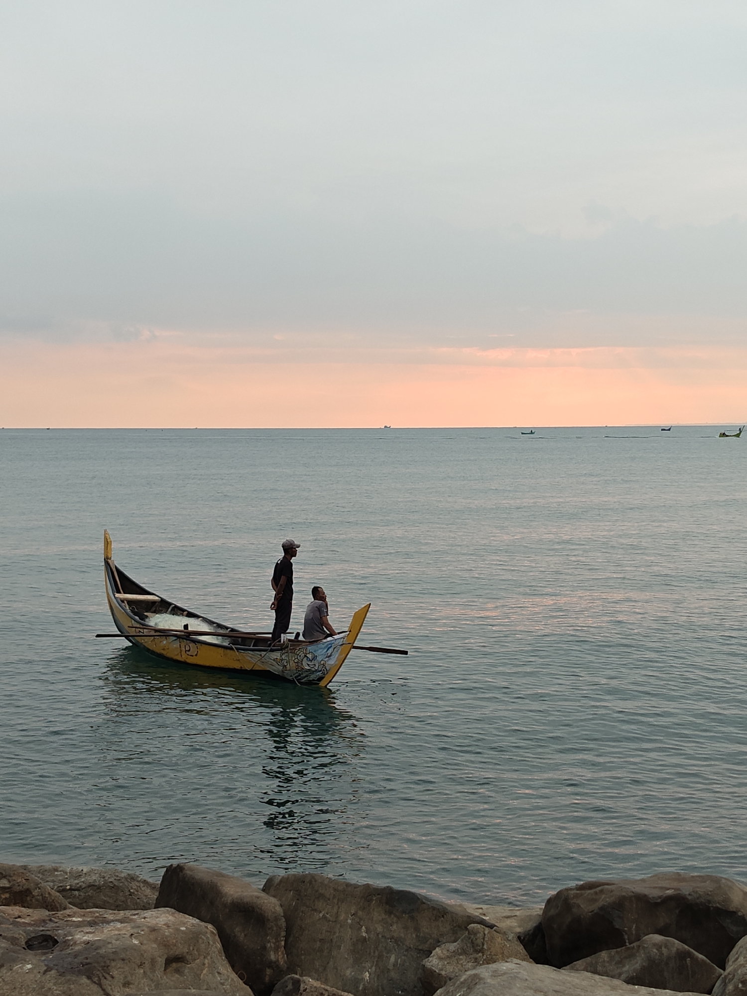 The image displays a coastal scene in Lhokseumawe, Indonesia, during either dawn or dusk, indicated by the orange-hued horizon. A traditional wooden fishing boat, primarily yellow with a raised bow and stern, is positioned in the mid-ground on calm, blue-green water. Decorative white and blue patterns are visible on the boat's bow. Two adult males occupy the boat. One man stands centrally, facing towards the right with his hands clasped behind his back, wearing a dark shirt, dark pants, and a light-colored baseball cap. The second man is seated closer to the bow, also generally facing right, dressed in a light-colored t-shirt and dark pants. A coiled fishing net is discernible within the boat's interior, and at least one oar extends horizontally from the boat's left side. Several small, dark silhouettes of other boats or vessels are visible on the distant horizon line. The immediate foreground comprises large, irregular dark gray and brown rocks, likely forming a breakwater or natural shoreline. The sky transitions from a pale gray-blue overhead to a warm orange-peach gradient at the horizon.