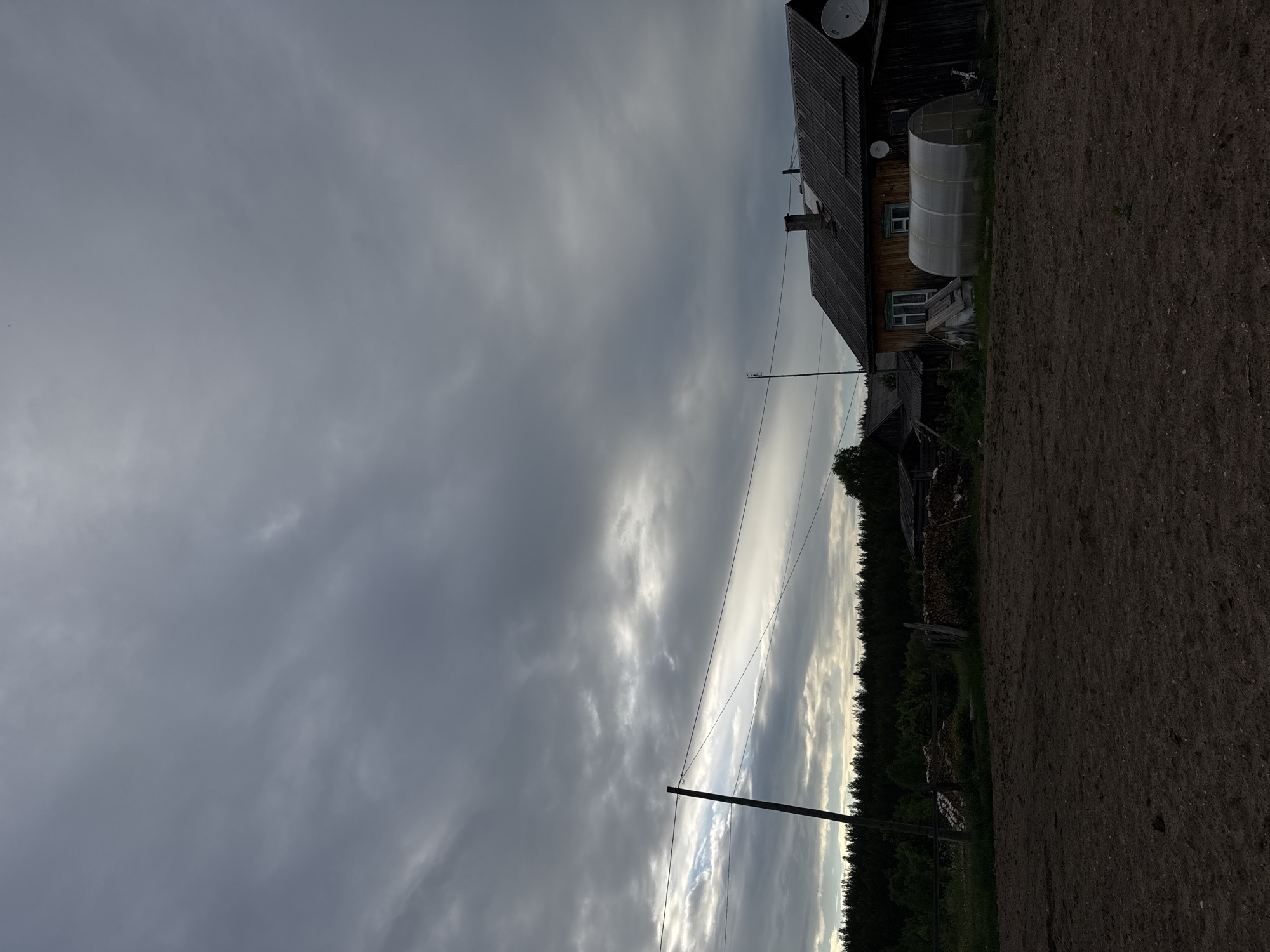 The image captures a rural outdoor scene in Ust'-Silayka, Russia, under a heavily overcast sky dominated by dense grey clouds, with slight lighter breaks visible towards the left horizon. In the lower third of the frame, a large area of dark brown, tilled soil is visible. A tall, dark wooden utility pole with multiple horizontal wires stands on the left side, extending into the frame. Behind the utility pole and spanning the mid-horizon is a dense, dark green treeline or forest. To the right, a wooden house with a dark, sloped roof, a visible chimney, and two white satellite dishes attached to its exterior is prominent. Two windows are discernable on the house's side. Directly in front of the house, a transparent, tunnel-shaped greenhouse is situated. Further back and slightly to the left of the main house, an additional dark wooden structure is partially visible, adjacent to a pile of stacked firewood. No people or animals are present. The lighting is dim and diffused, consistent with an overcast day.