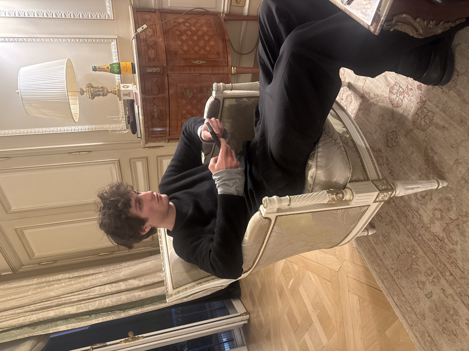 A young male with curly brown hair is seated in an ornate, classical-style armchair featuring a white, carved frame with gold accents and light-colored, patterned fabric upholstery. He wears a dark sweater over a grey long-sleeved shirt, black trousers, and dark shoes. Both hands hold a black smartphone, and his gaze is directed off-camera to the right. The floor consists of light-colored parquet wood, partially covered by a patterned oriental rug. To the viewer's left, a tall French window, framed by light-colored patterned curtains, reveals a dark exterior with a wrought-iron balcony railing. To the right, a polished wooden commode with marquetry detailing and brass hardware supports a table lamp with a pleated white shade and an ornate gold-colored base. A bottle of Veuve Clicquot champagne stands next to the lamp, alongside other indistinct objects. The walls are light-colored with decorative moldings and paneling. The scene depicts an elegant interior, consistent with a high-end residence or hotel suite in Paris, France.