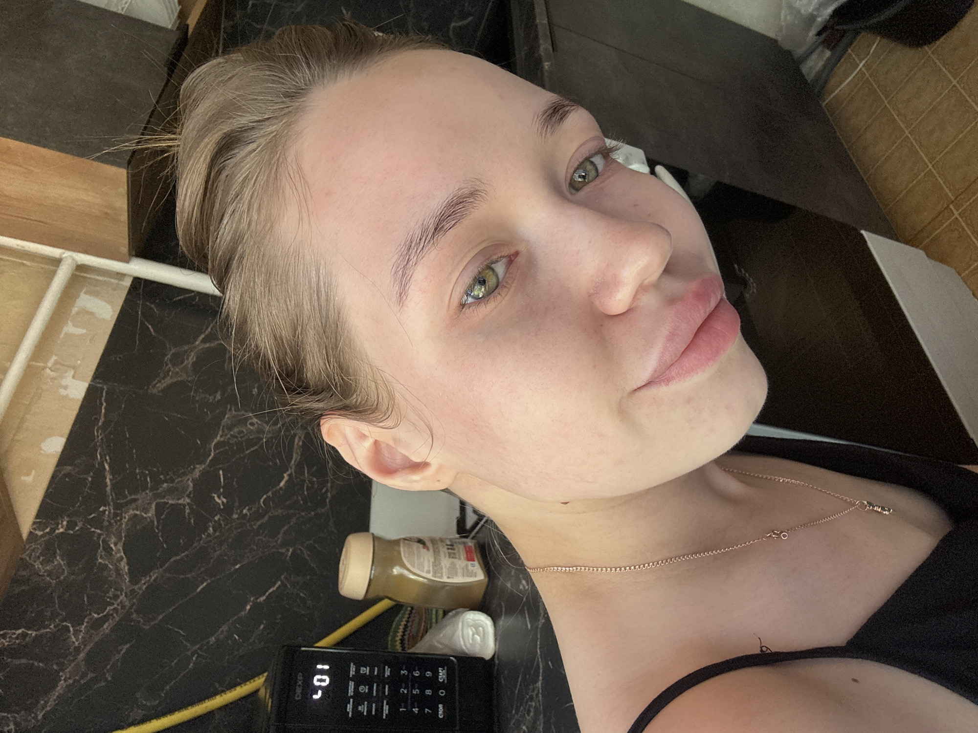 A selfie of a woman with fair skin and light brown hair pulled back, wearing a thin gold chain necklace and a black tank top. Her eyes are green. The background includes a black marble-patterned countertop, a microwave displaying "10," a jar of nut butter, and assorted kitchen items. The location appears to be an indoor kitchen environment. There is no verifiable evidence of Chaikovsky, Russia.