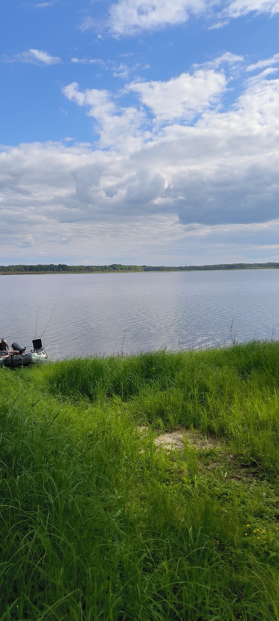 The image depicts a serene lake scene under a partly cloudy sky. In the foreground, lush green grass grows along the bank. To the left, an inflatable boat is moored near the shore, equipped with fishing rods and two outboard motors. The lake stretches out towards a distant, tree-lined horizon. The scene is quiet and appears to be a natural landscape, possibly in the city of Kurgan, Russia, known for its numerous lakes and natural beauty.