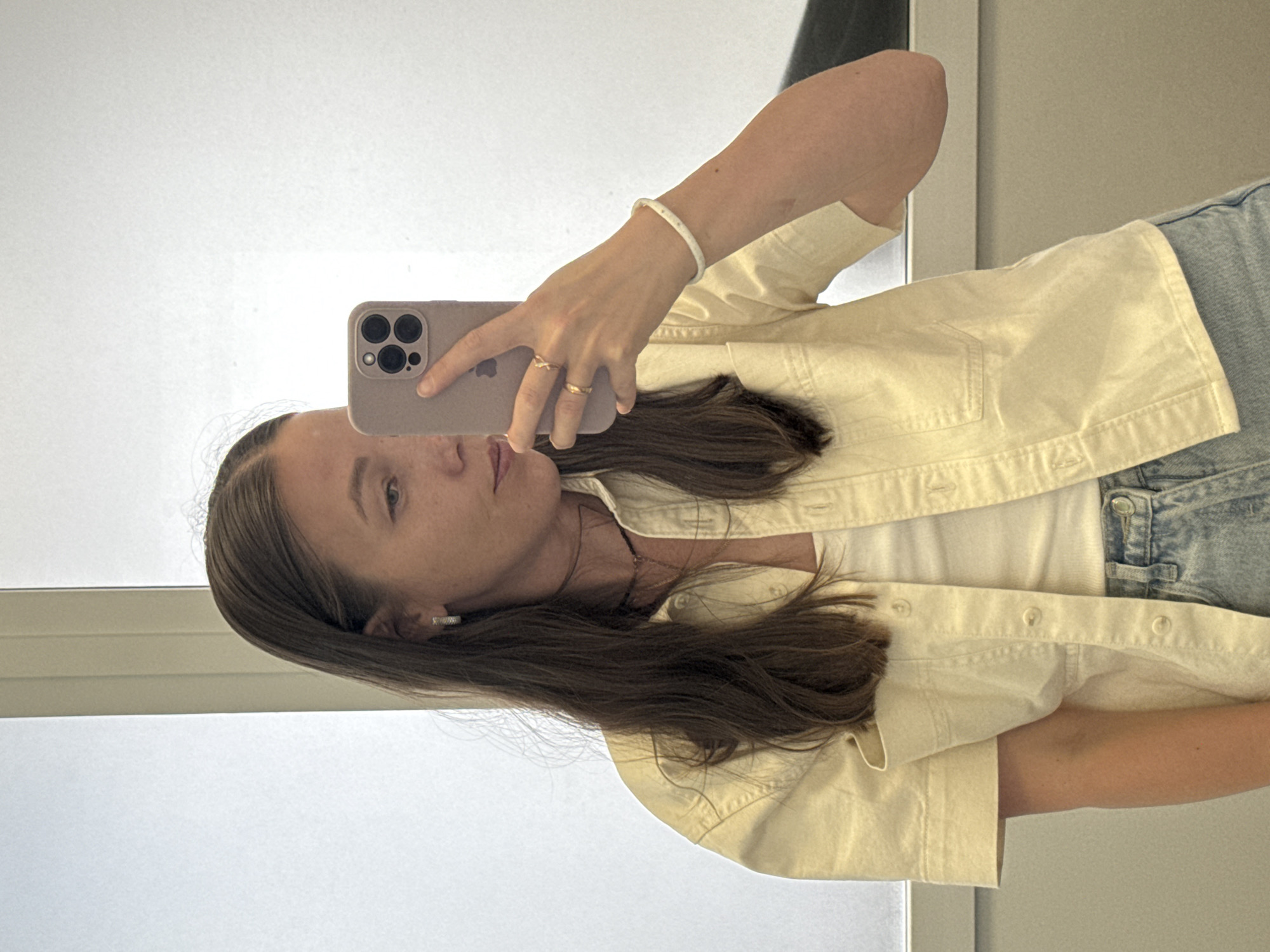 A woman with long, dark brown hair is taking a mirror selfie in what appears to be a fitting room or a room with bright, diffused lighting. She is wearing a light yellow button-down shirt with short sleeves, open to reveal a white tank top underneath. Her lower half is clad in light-wash denim jeans. She is wearing small hoop earrings and several rings on her left hand, which is holding a light purple smartphone. A white beaded bracelet adorns her left wrist. The background is a plain, light-colored wall with vertical lines, possibly a window or partition. There is no indication of the city of Chaikovsky, Russia, or any specific urban environment in the image.