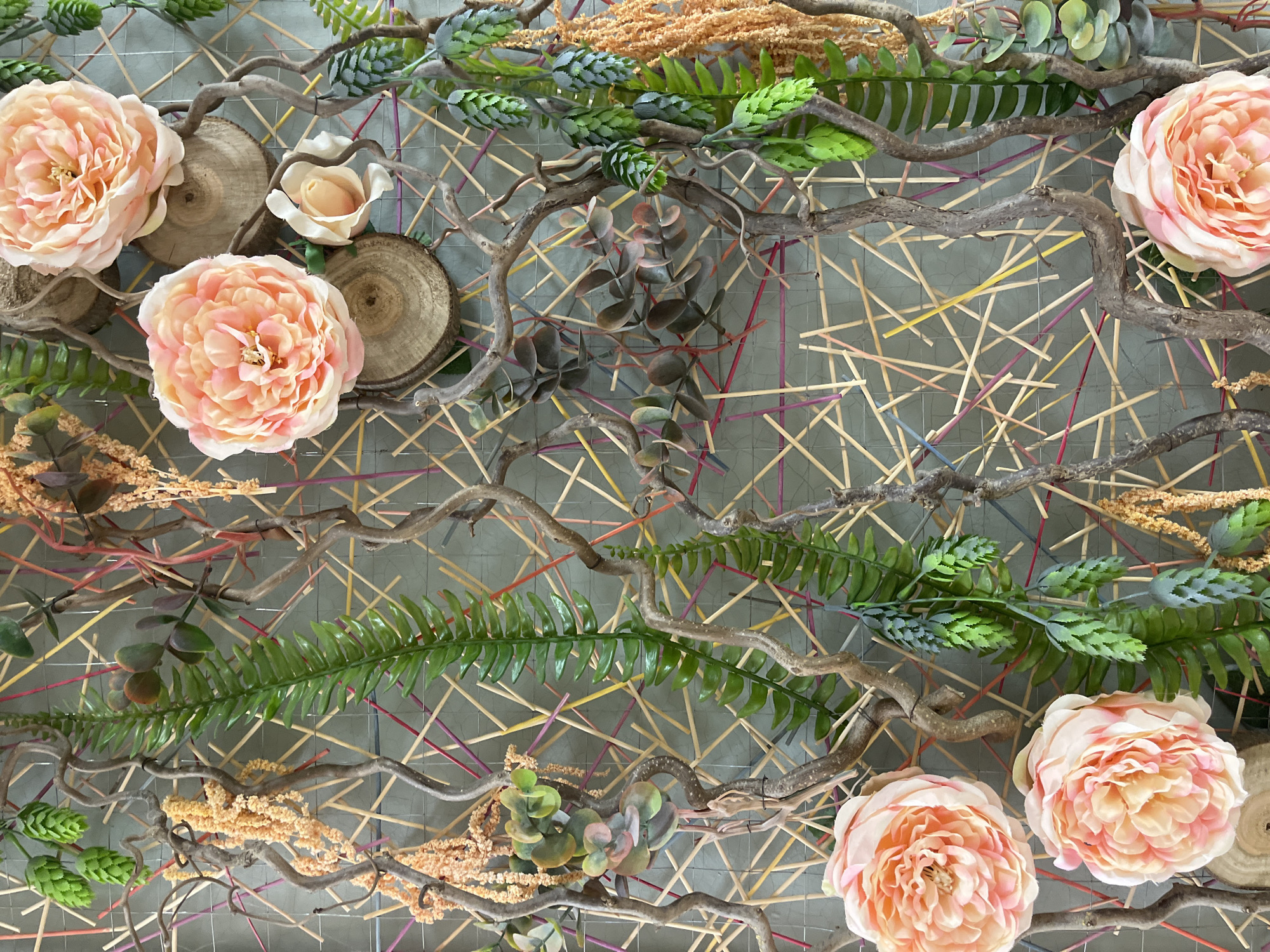 The image shows a decorative floral arrangement against a geometric backdrop. The backdrop consists of a wire grid with colored wooden sticks arranged diagonally within the grid. The floral arrangement includes several light pink roses, branches, green ferns, and small wooden discs. The arrangement is positioned in the foreground, obscuring parts of the grid. The image does not provide any information about its location in Saint Petersburg, Russia, or include details about people, actions, or interactions.