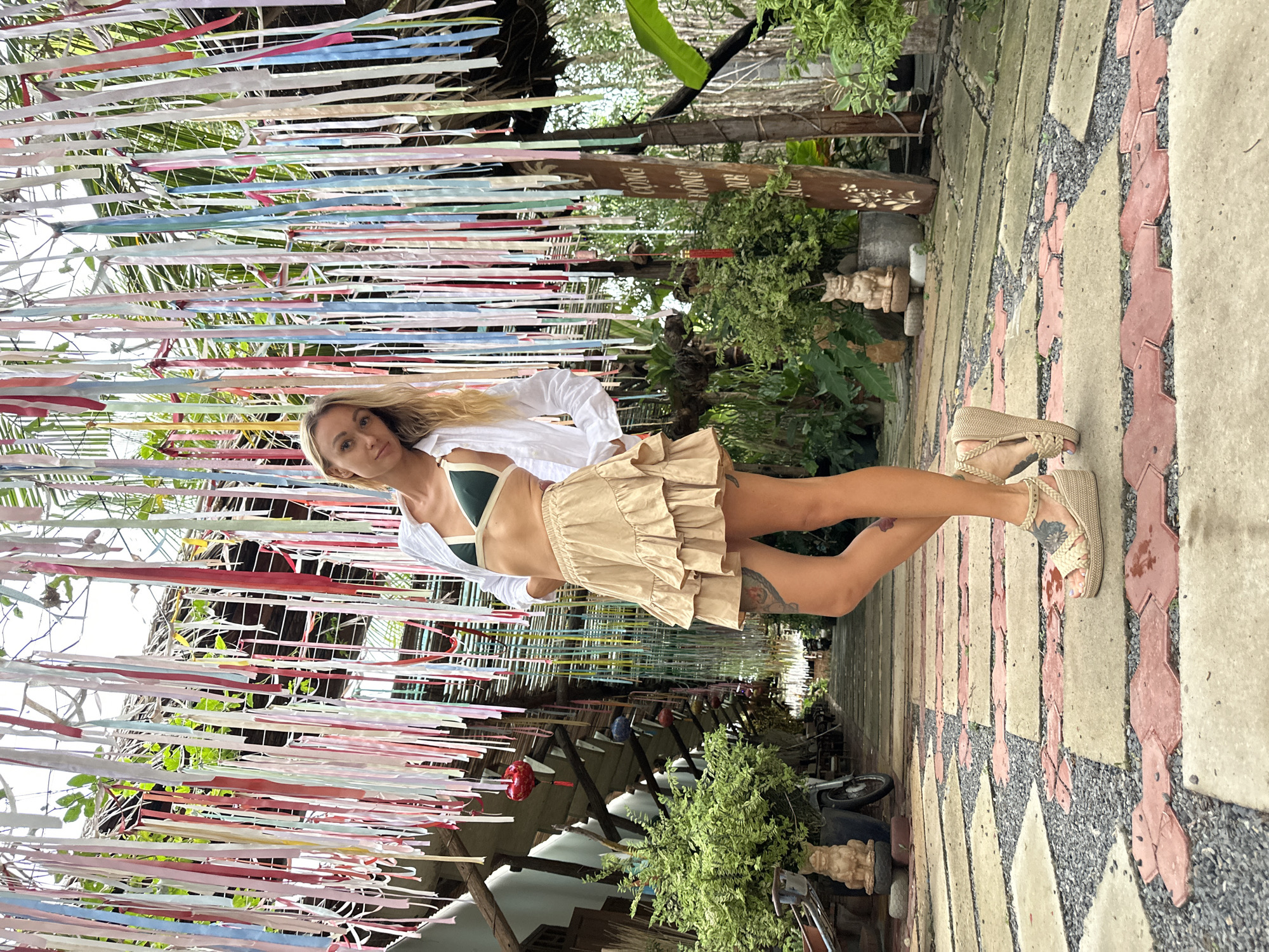 A young, fair-skinned woman is standing on a paved walkway in Mũi Né, Vietnam. She is wearing a dark green bikini top, a short beige ruffle skirt, and a white button-down shirt. Her feet are adorned with beige braided wedge sandals, and tattoos are visible on her legs. She is gazing towards the camera, her blonde hair hanging down.
Above her, a canopy of colorful ribbons stretches overhead. The walkway under her feet features a geometric pattern, with light gray tiles accented by insets of pink and black gravel. Various potted plants and greenery flank the path, including a small statue and a vintage motorcycle. A wooden sign with Vietnamese characters is visible behind the plants.