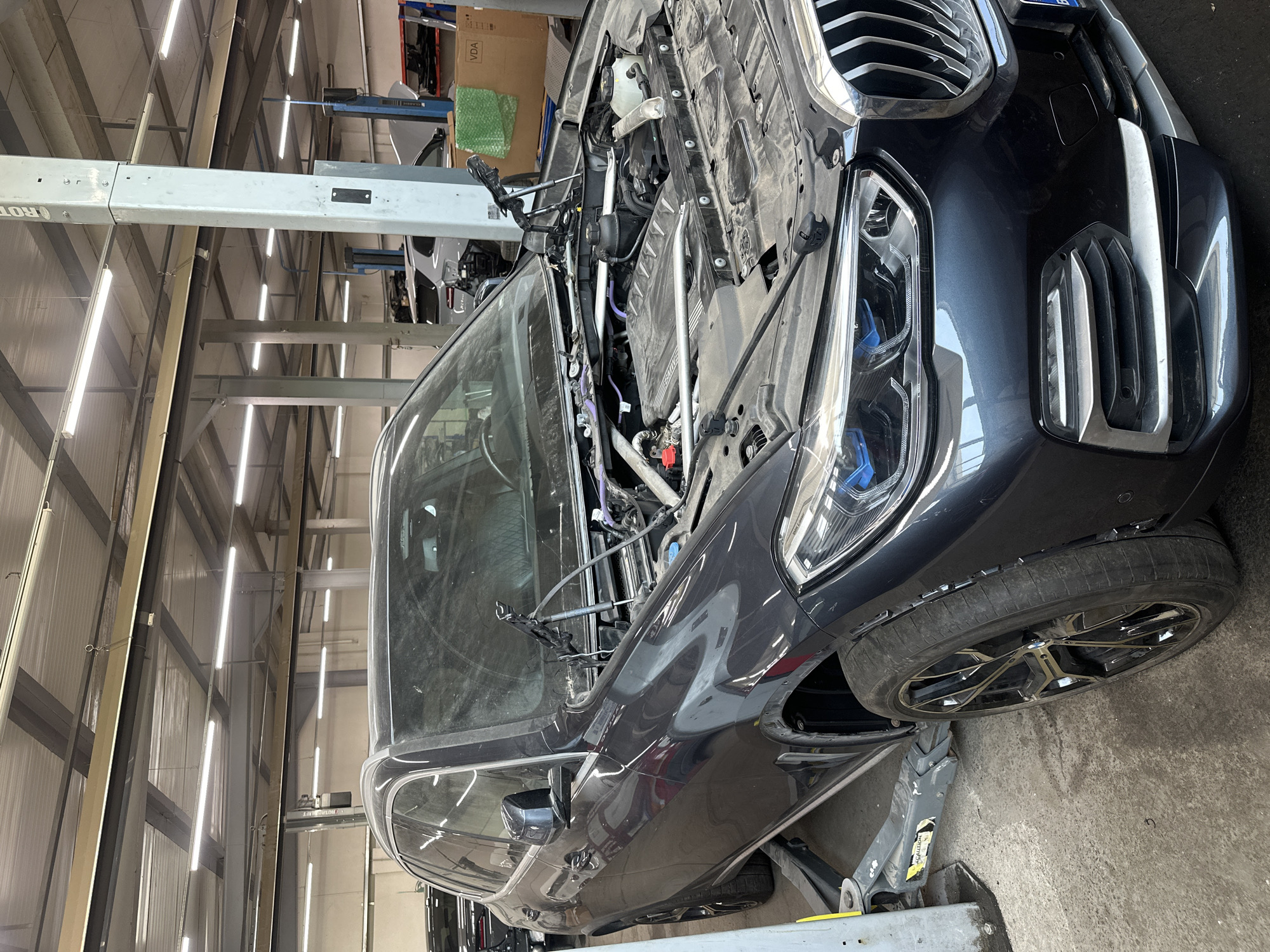 A dark-colored, possibly dark grey or black, BMW SUV or crossover vehicle is positioned in what appears to be an automotive repair shop or garage. The vehicle's hood is wide open, revealing the engine bay, which contains various components, wires, and hoses, some of which are purple. The front bumper is either partially removed or in a state of disassembly, with the intricate grille and modern LED-style headlights clearly visible. A blue detail is noticeable within the headlights, possibly indicating specific technology. The front passenger wheel, bearing a "BMW" logo on its hub and a stylish multi-spoke design, is visible, and the car's front end is supported by a robust grey floor jack with a yellow "CAUTION" sticker.
The setting is an indoor workshop characterized by high ceilings, exposed metal support beams, and bright, long fluorescent strip lights that illuminate the space. In the background, sections of white or light-colored walls are visible, along with shelves holding cardboard boxes, one of which has "VDA" printed on it. A car lift, a common piece of garage equipment, is partially visible with its grey metal structure. No people are present in the image, and the overall scene suggests active maintenance or repair work being performed on the vehicle. The original image is rotated 90 degrees clockwise, causing the car to appear on its side.
