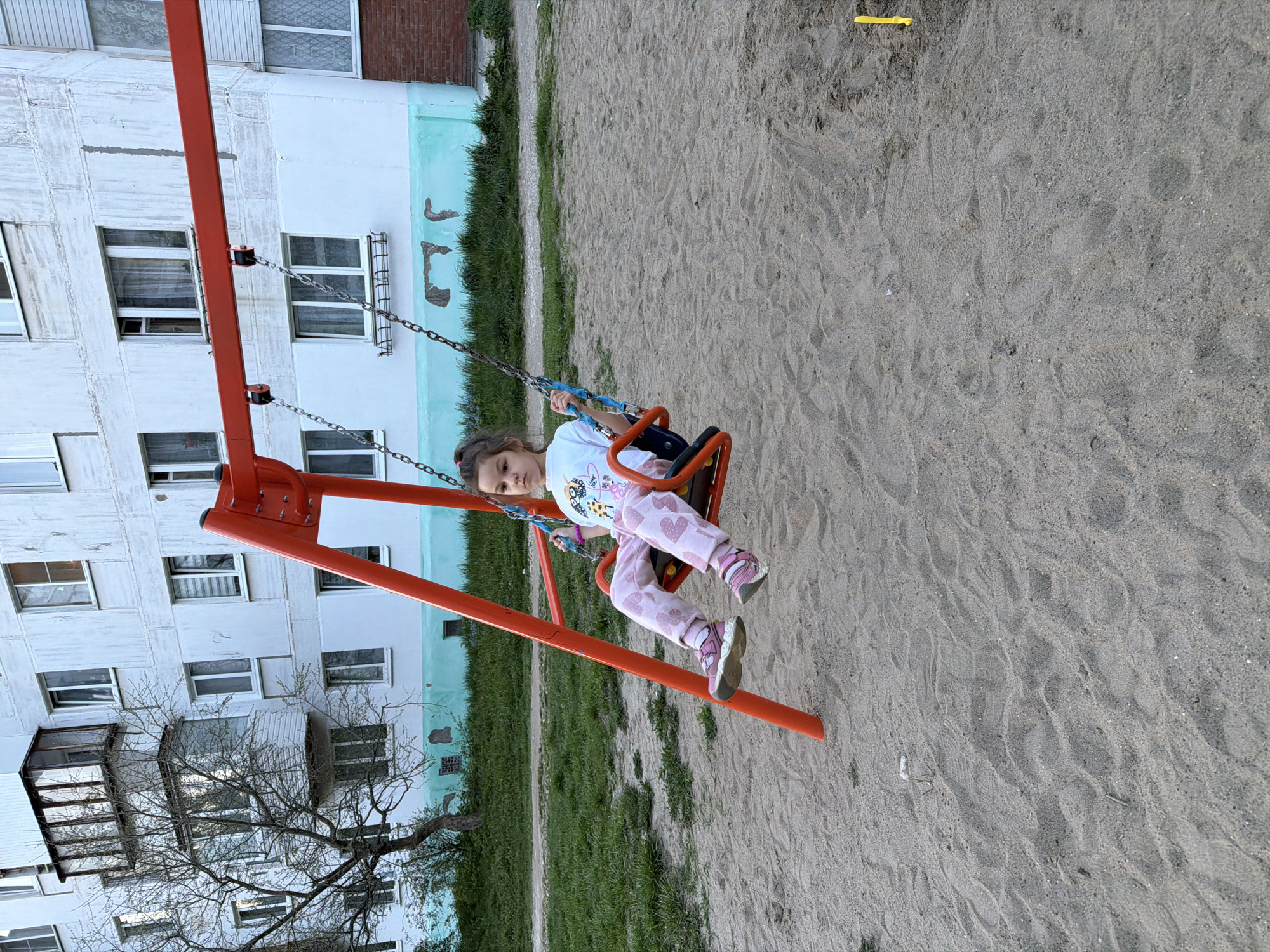 A young child is sitting on a swing at a playground. The swing has a red frame and a blue seat. The child is wearing a white shirt with a graphic print and pink pants with a heart pattern. She is holding the swing chains with her hands. The playground surface appears to be sand. In the background is a multi-story residential building with visible windows and balconies. The lower section of the building is painted with a light blue trim. The image was taken in Chaikovsky, Russia.