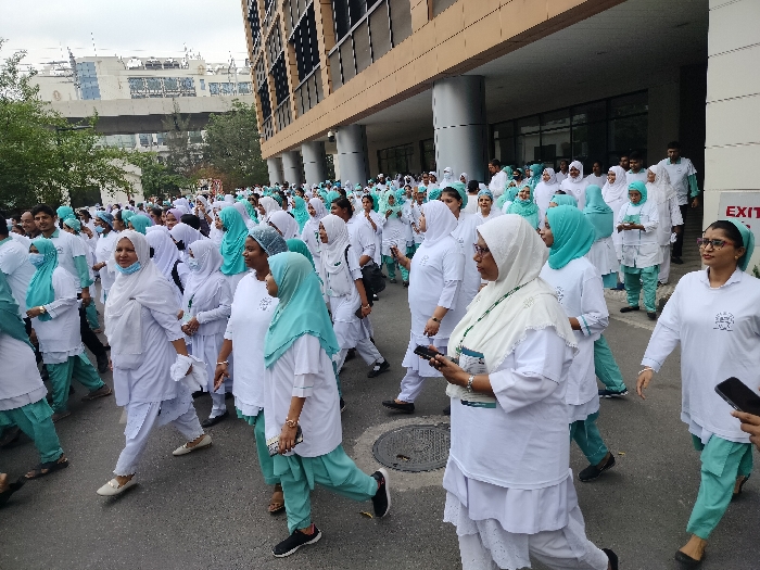 A large group of women, primarily wearing white tunics and light blue pants, walk on a paved street in front of a large, light-colored building. Some women are wearing white headscarves and a few wear light blue headscarves. They are walking in a single file line, and some are holding cell phones or other objects. The building is in the background, and there is a sign that reads "EXIT" on it. The scene is located in Dhaka, Bangladesh, at the coordinates 23.741, 90.395.