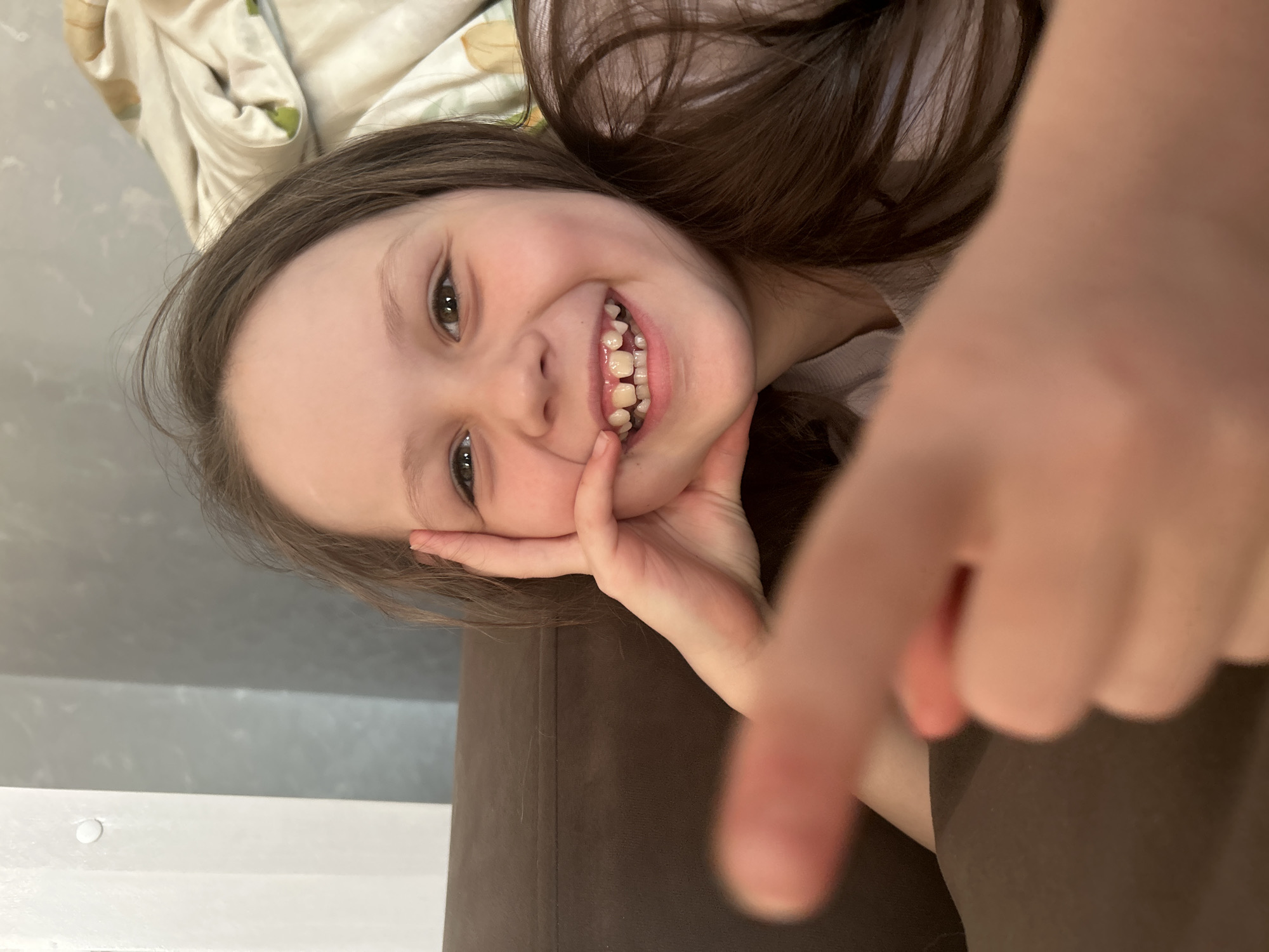 A young girl is lying on a brown couch, pointing with her right index finger towards the camera. She has light skin, brown hair, and is smiling, revealing her teeth. Her left hand is resting against her cheek. A light-colored patterned blanket is visible in the upper left corner. The location cannot be determined as Chaikovsky, Russia from this image.